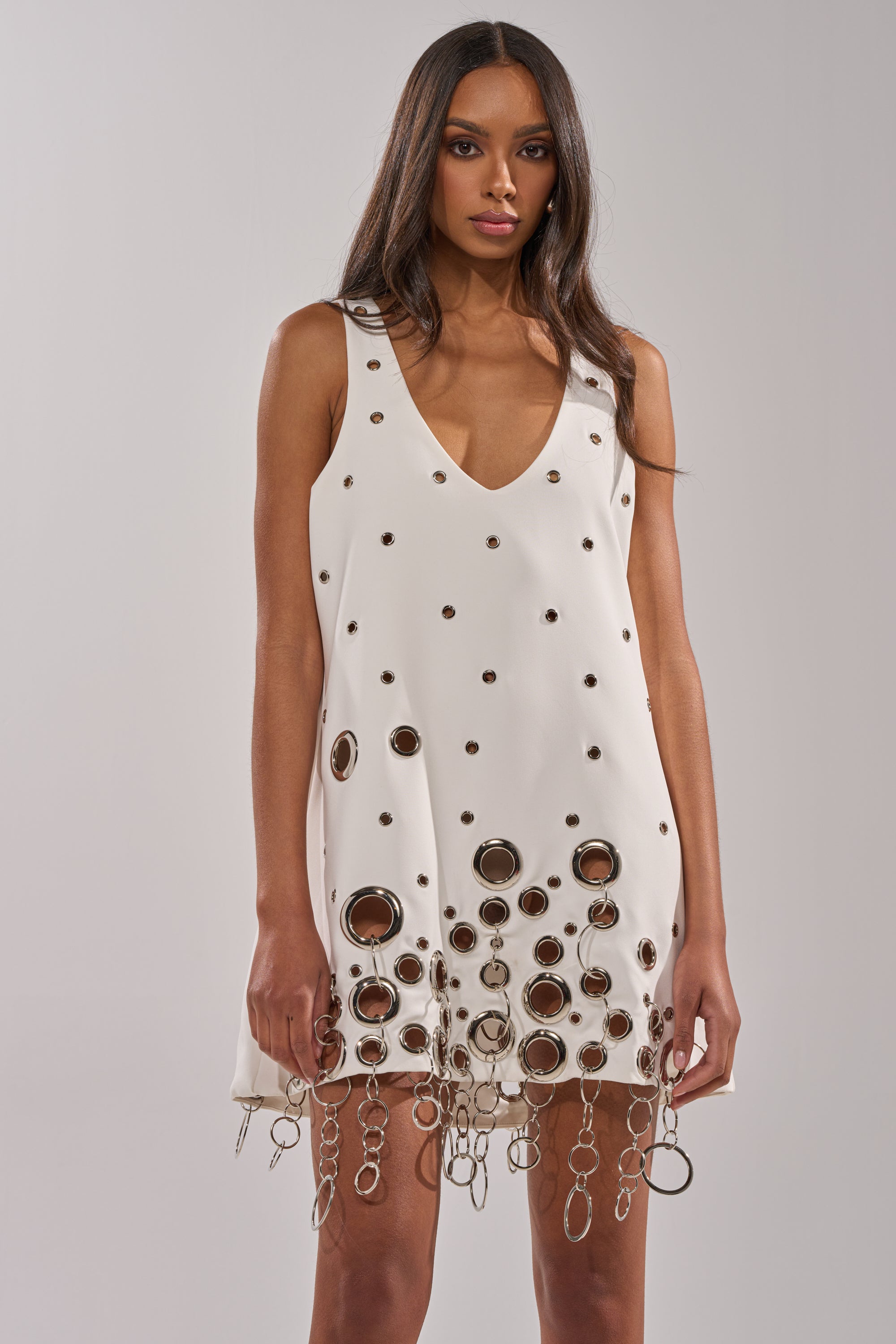 A woman wears the ONE OF A KIND MINI DRESS WITH METAL HARDWARE IN WHITE, featuring a deep V-neckline, metallic grommets, and chain fringe at the hem. She has long straight hair and stands against a plain background.