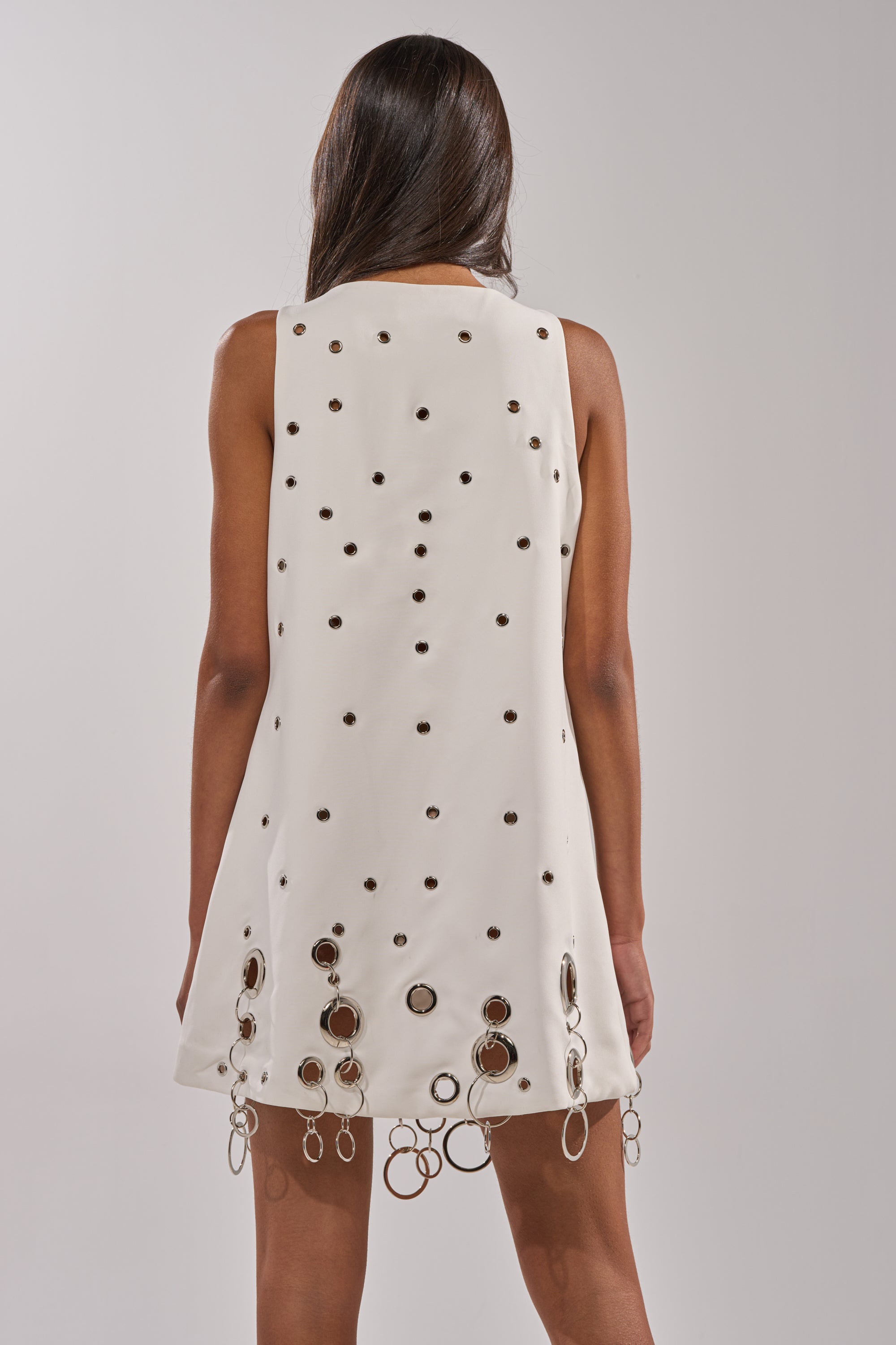 A woman faces away, wearing the ONE OF A KIND MINI DRESS WITH METAL HARDWARE IN WHITE, featuring metallic grommets, ring details, and chain fringe accents at the hem. Her straight dark hair falls loosely down her back.