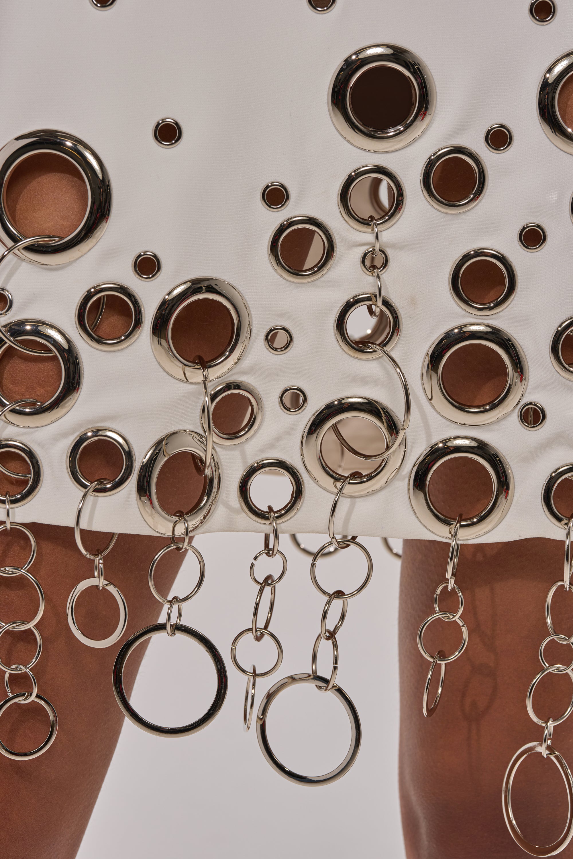 Close-up of the ONE OF A KIND MINI DRESS WITH METAL HARDWARE IN WHITE, featuring metallic rings, grommets, and chain links. Brown skin is visible through the dress’s cut-out details and embellishments.