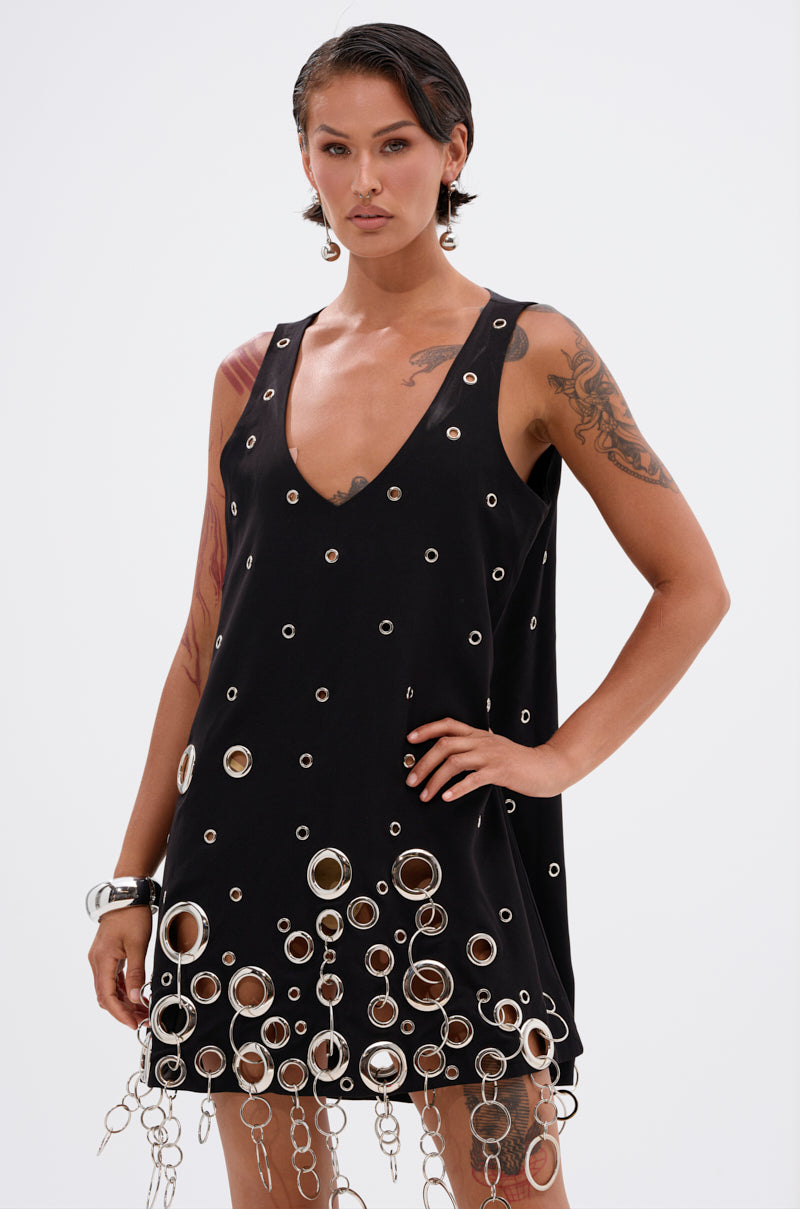 Woman with short dark hair stands confidently against a plain light background, wearing the ONE OF A KIND MINI DRESS WITH METAL HARDWARE—a sleeveless black dress adorned with unique metal details and rings.