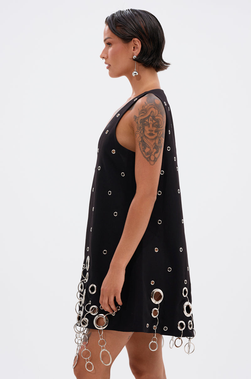 A person with short dark hair and a visible arm tattoo models the ONE OF A KIND MINI DRESS WITH METAL HARDWARE—featuring silver grommets and large hanging rings—with dangling earrings, against a plain light background.