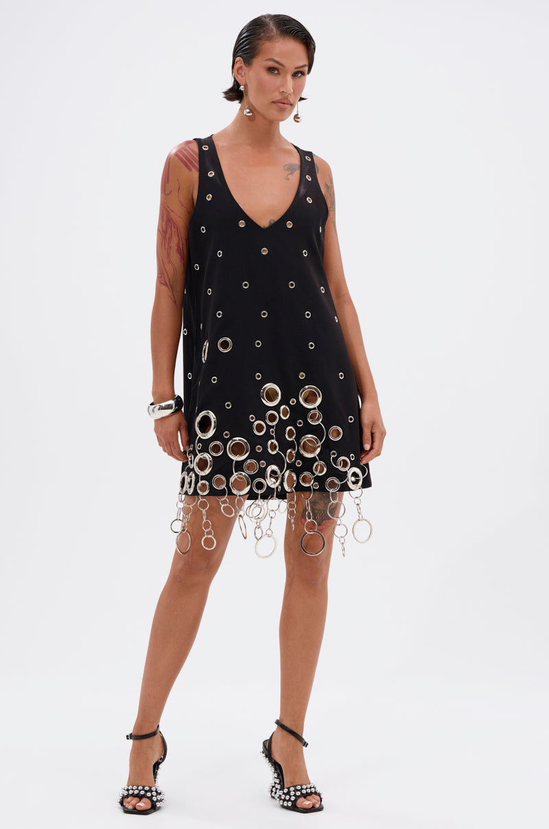 A woman in the ONE OF A KIND MINI DRESS WITH METAL HARDWARE—sleeveless, black, with silver grommets and dangling rings at the hem—pairs it with studded high-heeled sandals, bracelets, slicked-back hair, and visible tattoos.