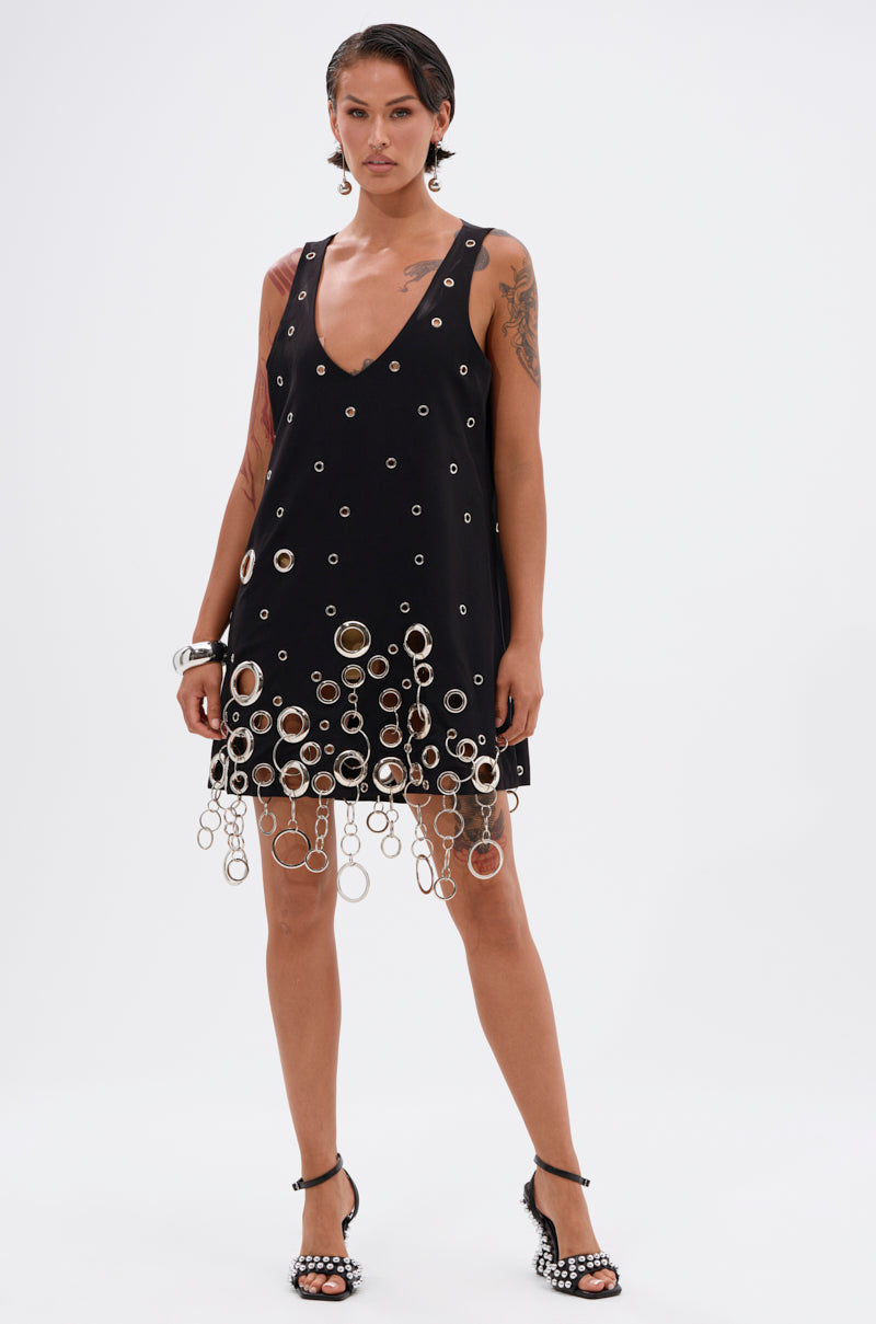A woman with short hair and visible tattoos poses confidently against a white background, wearing the ONE OF A KIND MINI DRESS WITH METAL HARDWARE and black high-heeled sandals with studs.