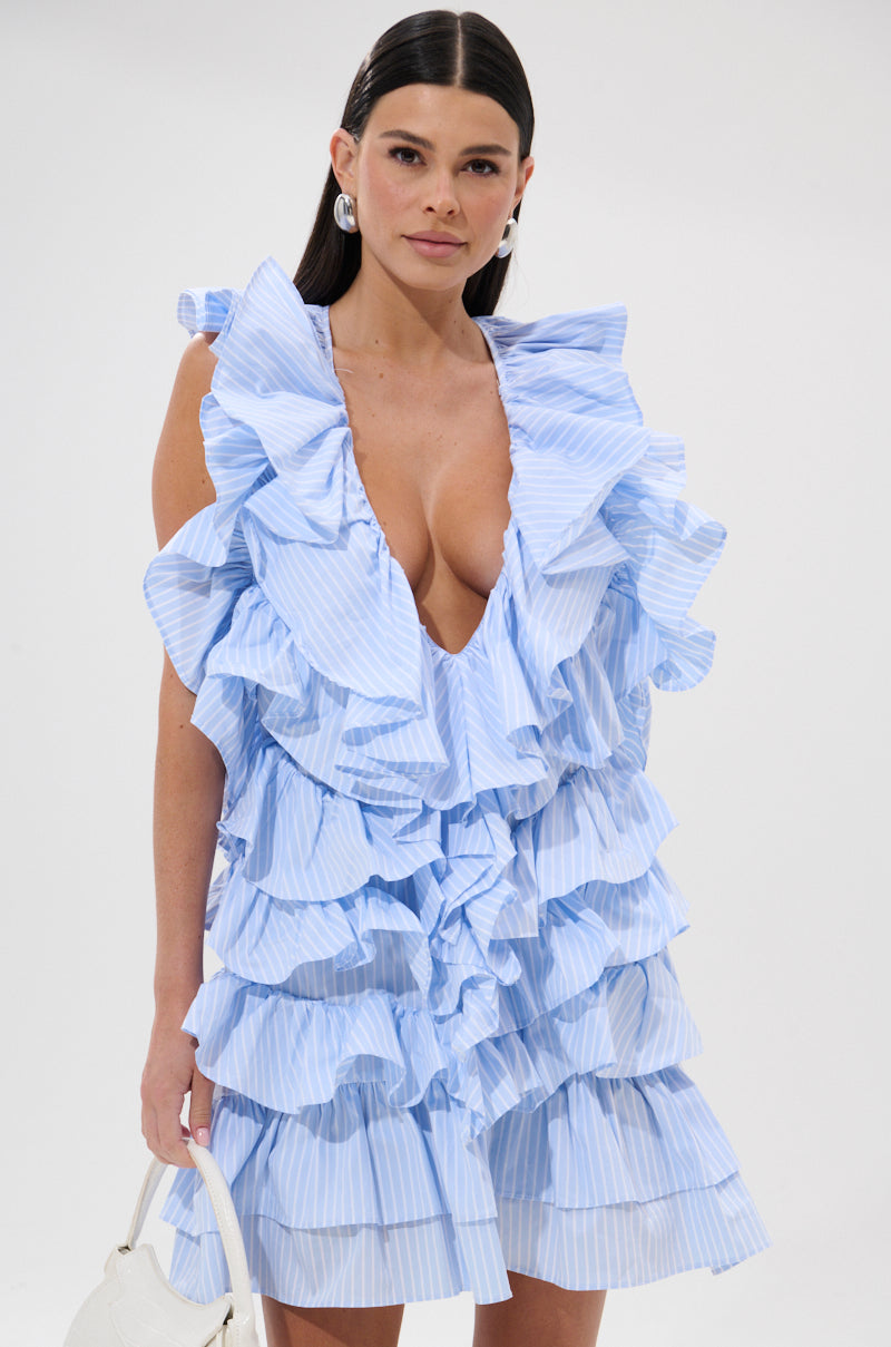 A woman with long dark hair models the ONCE UPON A TIME RUFFLED MINI DRESS, a sleeveless light blue mini dress with a deep V-neckline. She holds a white handbag and wears silver hoop earrings against a plain white background. 2