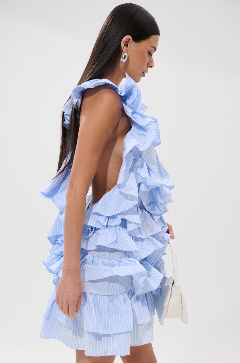 A woman with long dark hair models the ONCE UPON A TIME RUFFLED MINI DRESS, a light blue and white pinstripe style with ruffles and open sides, paired with large earrings and a small white handbag against a plain white background. 3