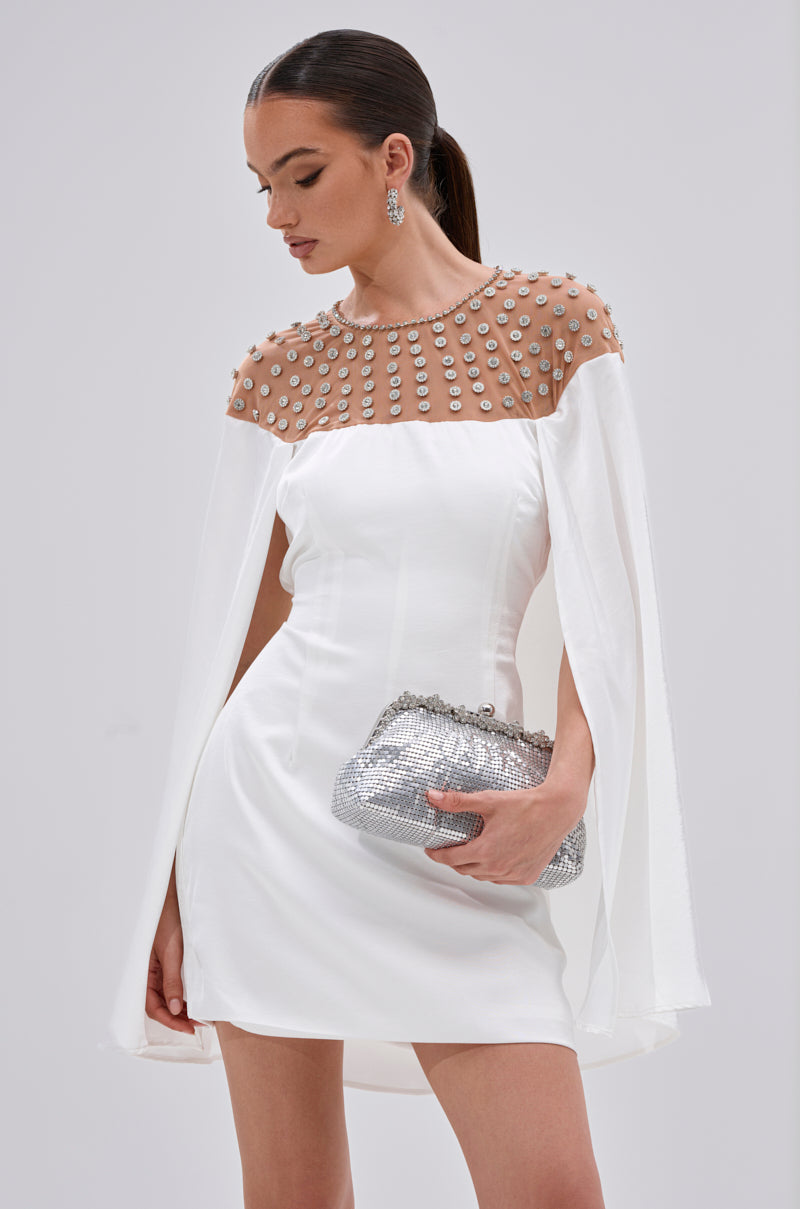 Perfect for cocktail events, she stuns in the LILA SATIN MINI DRESS WITH RHINESTONES, featuring a sheer beaded neckline and cape sleeves. A sparkling silver clutch and rhinestone earrings complete her elegant look as she gazes downward thoughtfully.