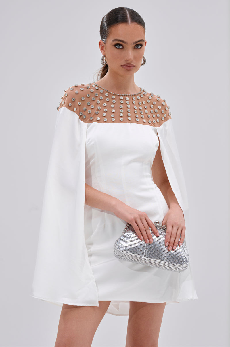 Wearing the LILA SATIN MINI DRESS WITH RHINESTONES, a woman with sleek hair and statement earrings holds a silver clutch. Her sheer, pearl-embellished cape sleeves radiate elegance, making her perfect for cocktail events against a light background.