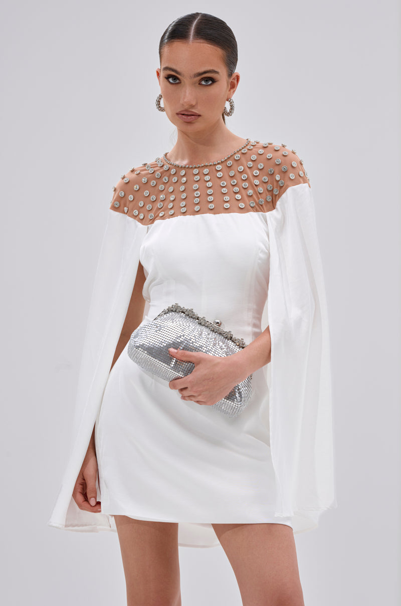 A woman wears the LILA SATIN MINI DRESS WITH RHINESTONES, featuring a sheer embellished neckline and cape sleeves. She pairs it with a silver clutch and hoop earrings, her hair sleek and pulled back—perfect for cocktail events.