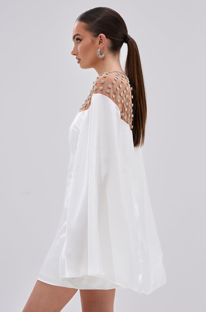 A woman stands in profile against a plain light background, wearing the LILA SATIN MINI DRESS WITH RHINESTONES and a sheer, pearl-embellished cape—an elegant look for cocktail events. Her straight brown hair is in a low ponytail, paired with large hoop earrings.