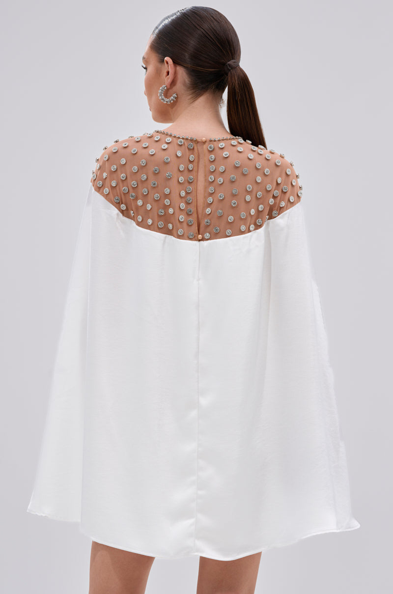 A woman with dark hair in a low ponytail is shown from the back wearing the LILA SATIN MINI DRESS WITH RHINESTONES, featuring a white satin finish and sheer rhinestone-embellished back panel. She pairs it with large hoop earrings for cocktail events.
