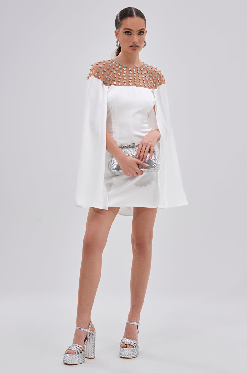 A woman wears the LILA SATIN MINI DRESS WITH RHINESTONES, featuring sheer, rhinestone-embellished shoulders and cape sleeves. She completes her chic cocktail look with a silver clutch, platform sandals, hoop earrings, and sleek pulled-back hair.