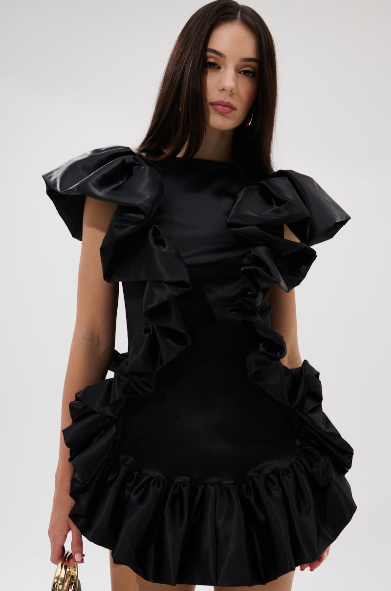 A woman with long, straight brown hair wears THE MOMENT IS MINE SATIN MINI DRESS, featuring curve-hugging satin fabric and dramatic ruffled details on the shoulders, front, and hem. She holds a small gold handbag against a plain white background.
