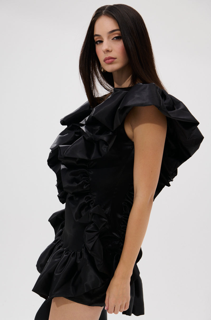 A woman with long dark hair poses against a white background, wearing THE MOMENT IS MINE SATIN MINI DRESS, a black mini dress with dramatic ruffles and a high neckline.