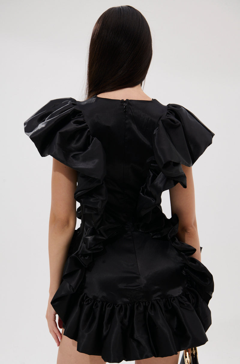 A woman with long dark hair is seen from behind wearing THE MOMENT IS MINE SATIN MINI DRESS—a curve-hugging black mini dress with dramatic ruffled sleeves and a ruffle hem, set against a plain light background.