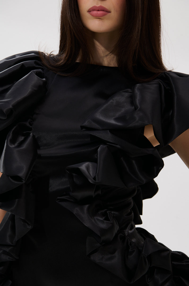 A woman wears THE MOMENT IS MINE SATIN MINI DRESS, a black satin mini with bold ruffled details on the shoulders and bodice. Her long dark hair is down, face partly visible against a light background—a chic look for cocktail parties.