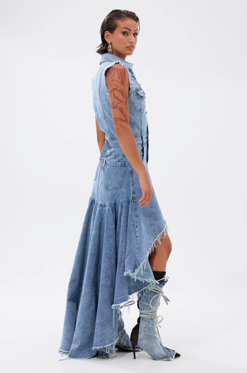 A person stands sideways in the READ IT AND WEEP DENIM MINI DRESS, featuring a light-wash finish, frayed train, and ruffled hem. They pair it with matching denim boots and show off a bold arm tattoo against a white background.