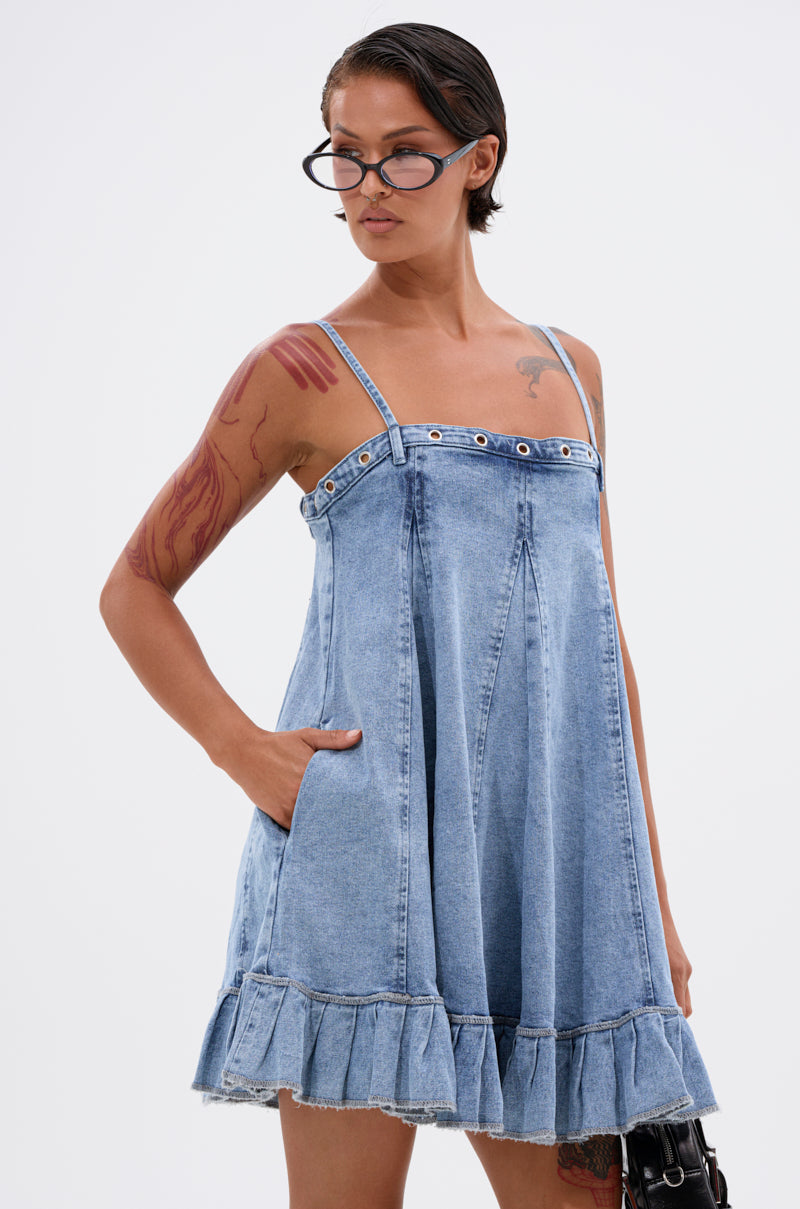 A person with short, slicked-back hair and glasses, showing arm tattoos and one hand in a pocket, wears the PLEATED UP MINI DRESS IN DENIM—a sleeveless mini dress with a ruffled hem—standing against a plain background. 0