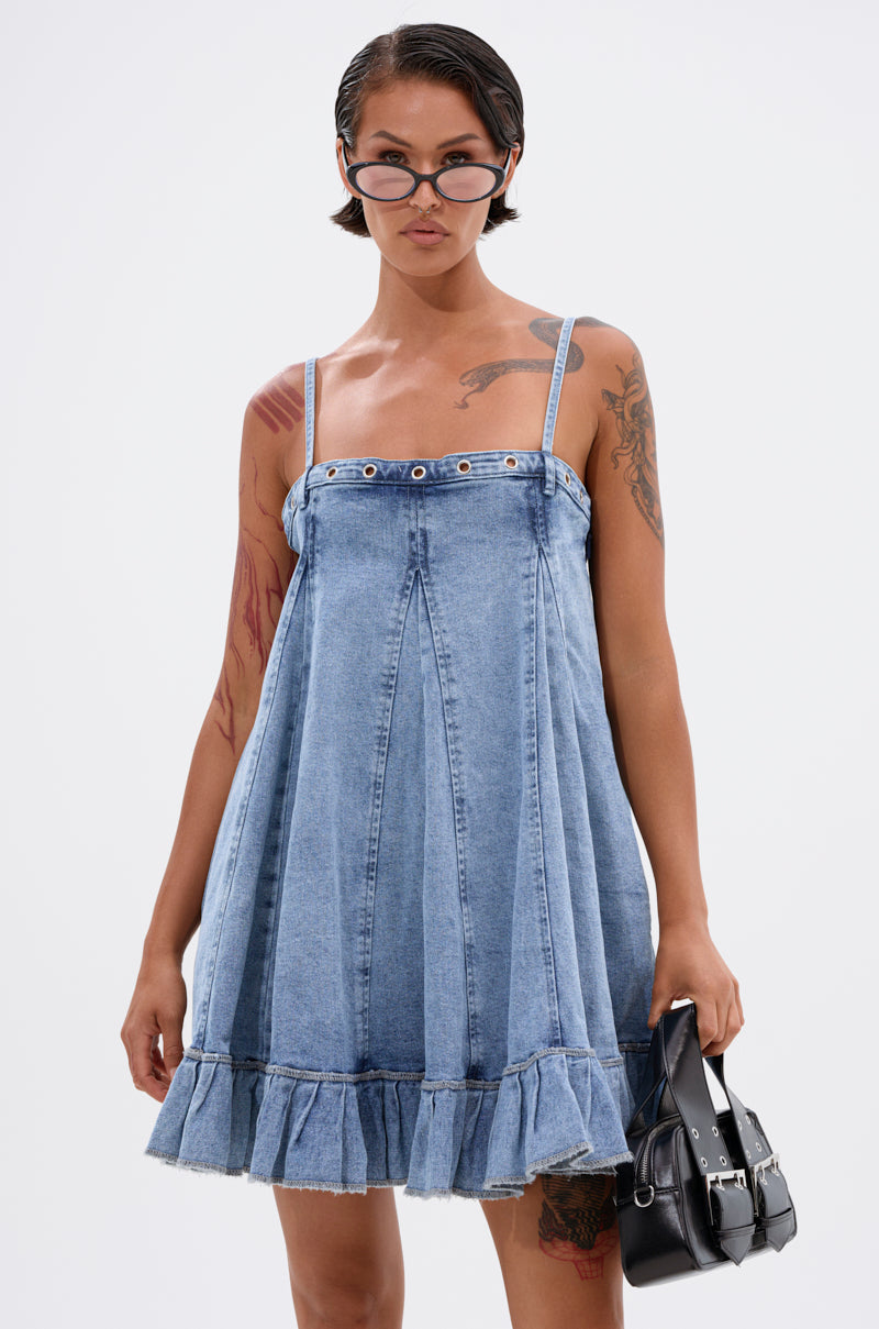A person with short dark hair wears the PLEATED UP MINI DRESS IN DENIM, featuring a ruffle hem. They style it with narrow sunglasses, a black handbag, and display tattoos on their arms and thighs. The background is plain white. 1