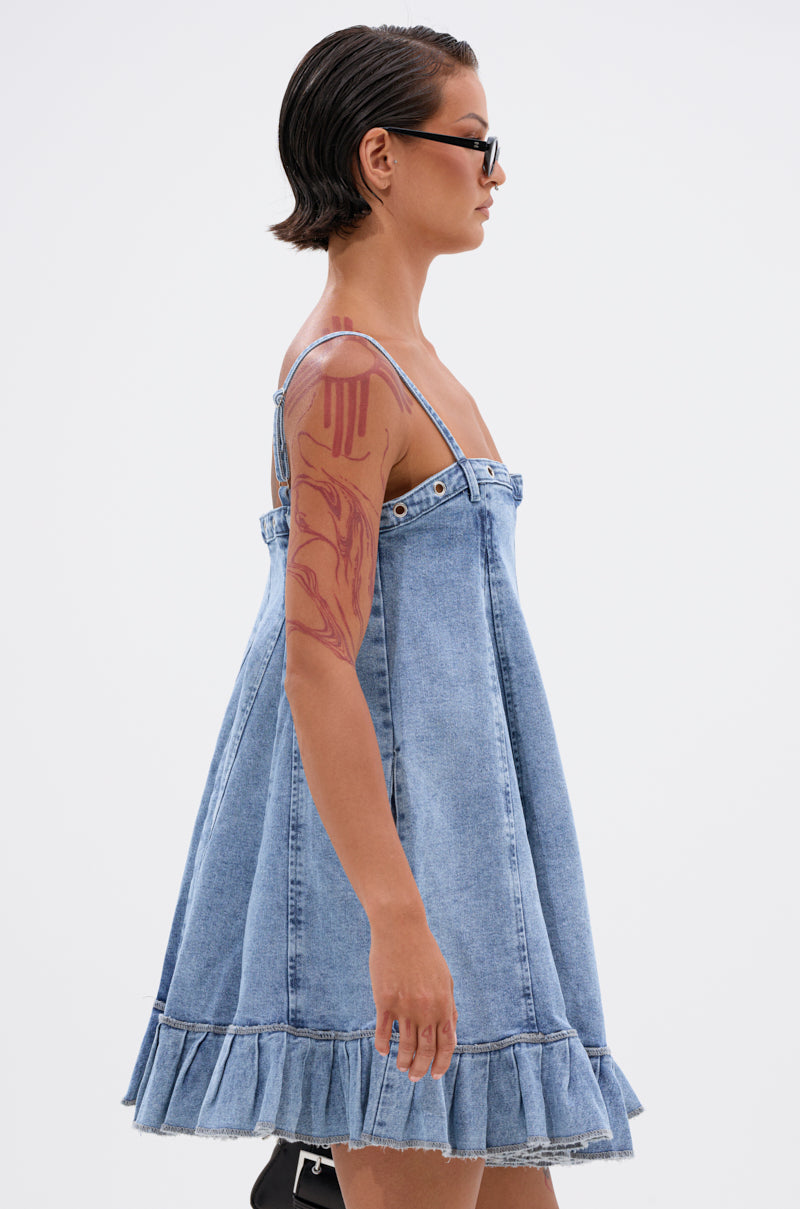 A person with short dark hair and sunglasses models the PLEATED UP MINI DRESS IN DENIM, a loose, sleeveless mini dress with a ruffle hem, showing a red upper arm tattoo against a plain white background. 3