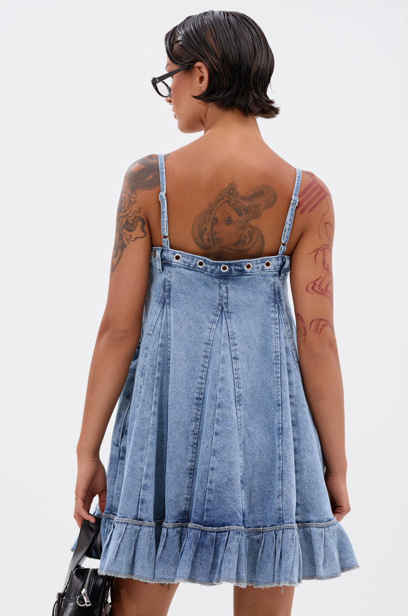 A person with short dark hair and visible back and arm tattoos is seen from behind holding a black handbag, wearing the PLEATED UP MINI DRESS IN DENIM—a sleeveless, flared mini dress with a ruffle hem. 4