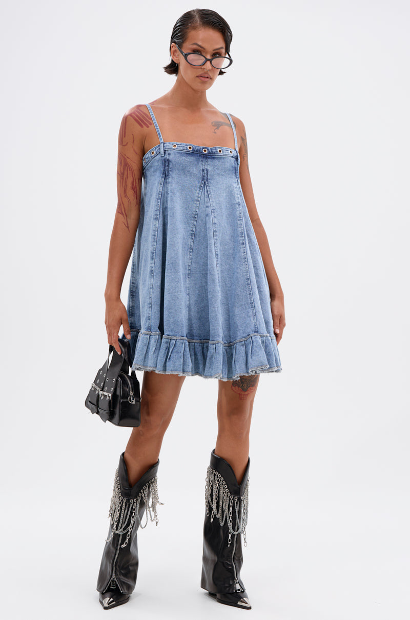 Wearing the PLEATED UP MINI DRESS IN DENIM, black pointy boots with silver chains, a small black handbag, glasses, and showing tattoos, a person stands against a plain white background. 5