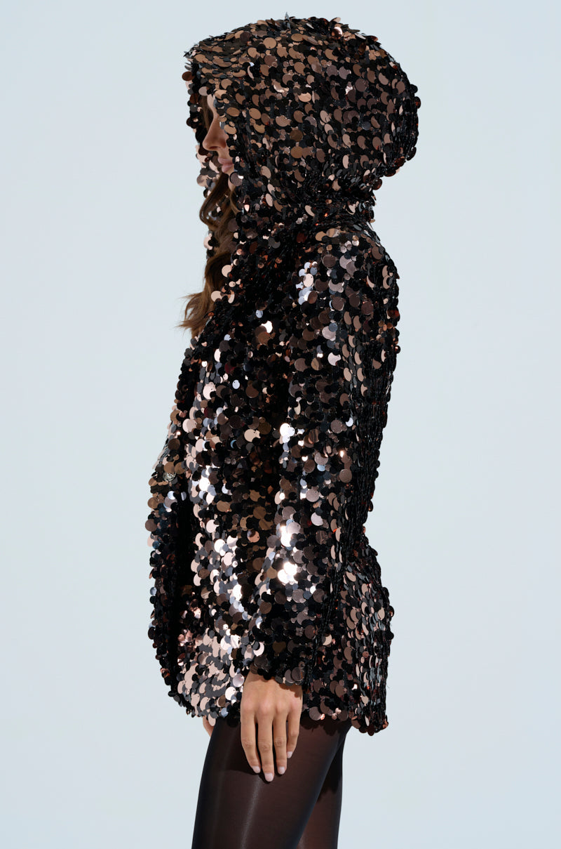 The person is wearing a shiny, large-circled sequined PARTY AT JOSH'S SEQUIN HOODED BLAZER, reflecting light. Paired with black leggings, the AKIRA-inspired outfit features the hood up as they stand facing sideways.