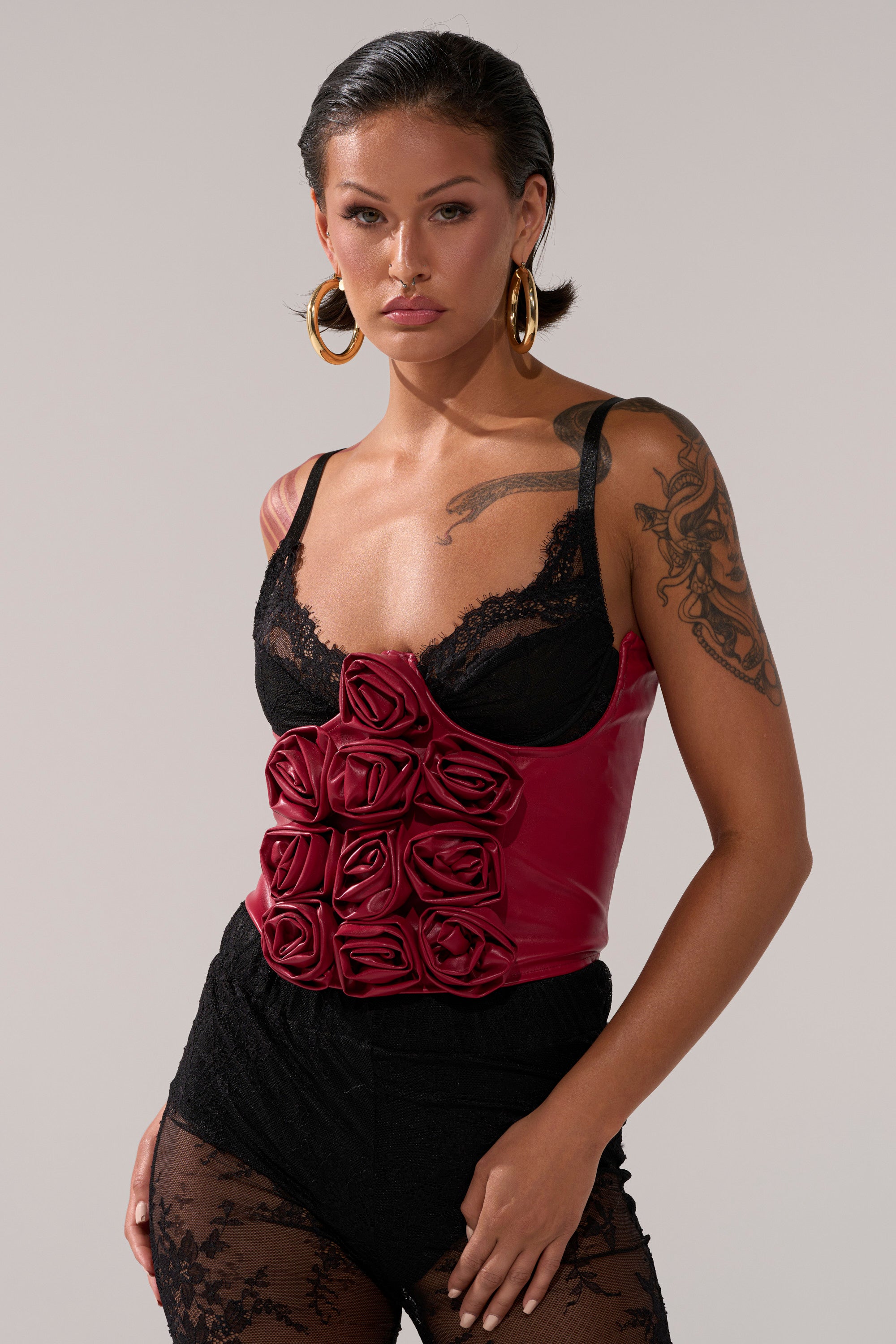 A woman with slicked back hair and large hoop earrings wears a black lace top, black lace shorts, and the striking IN BLOOM PU BELT adorned with rose-shaped details, standing confidently against a plain background.