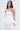 A woman with long, wavy brown hair models the IM TOO GOOD FLARE BELT, a strapless white corset top with a peplum silhouette and matching white pants, standing against a plain light background.