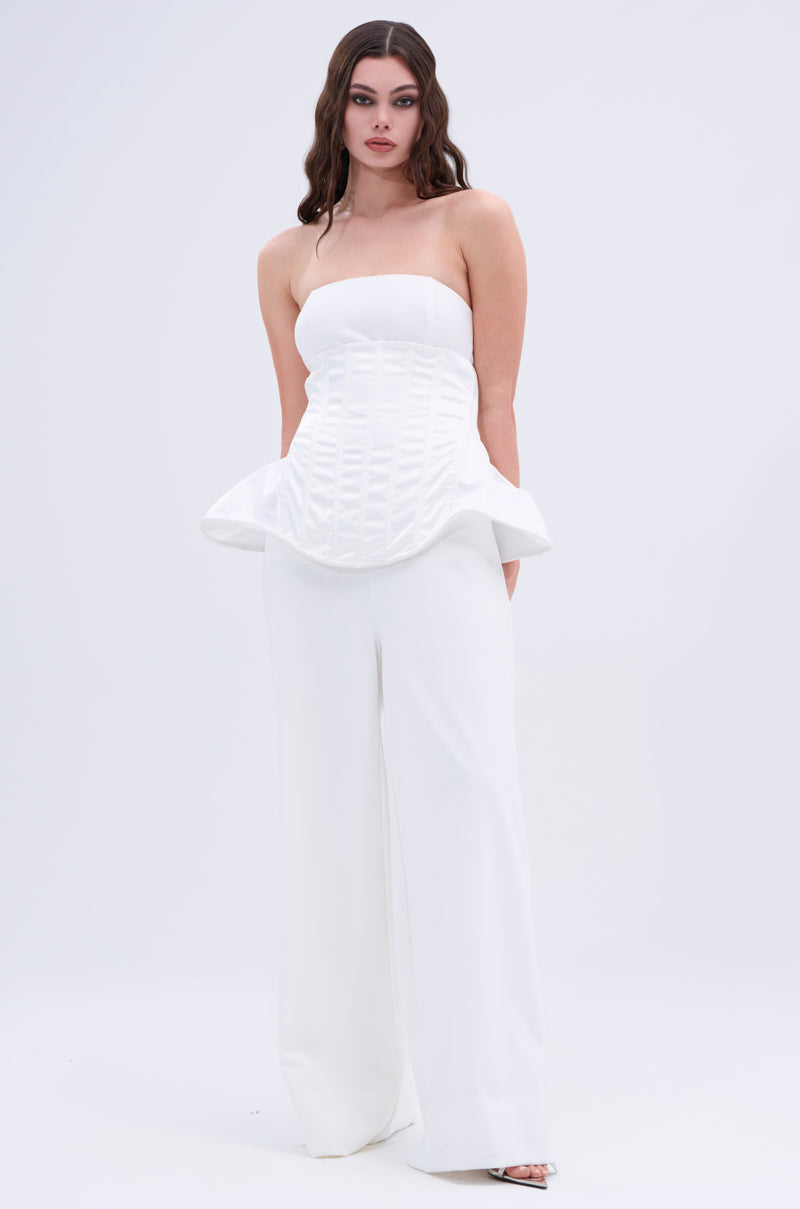 A woman with long wavy hair and a neutral expression stands against a plain background, wearing the IM TOO GOOD FLARE BELT—a strapless white structured peplum top with wide-leg white pants and open-toe heels. 1