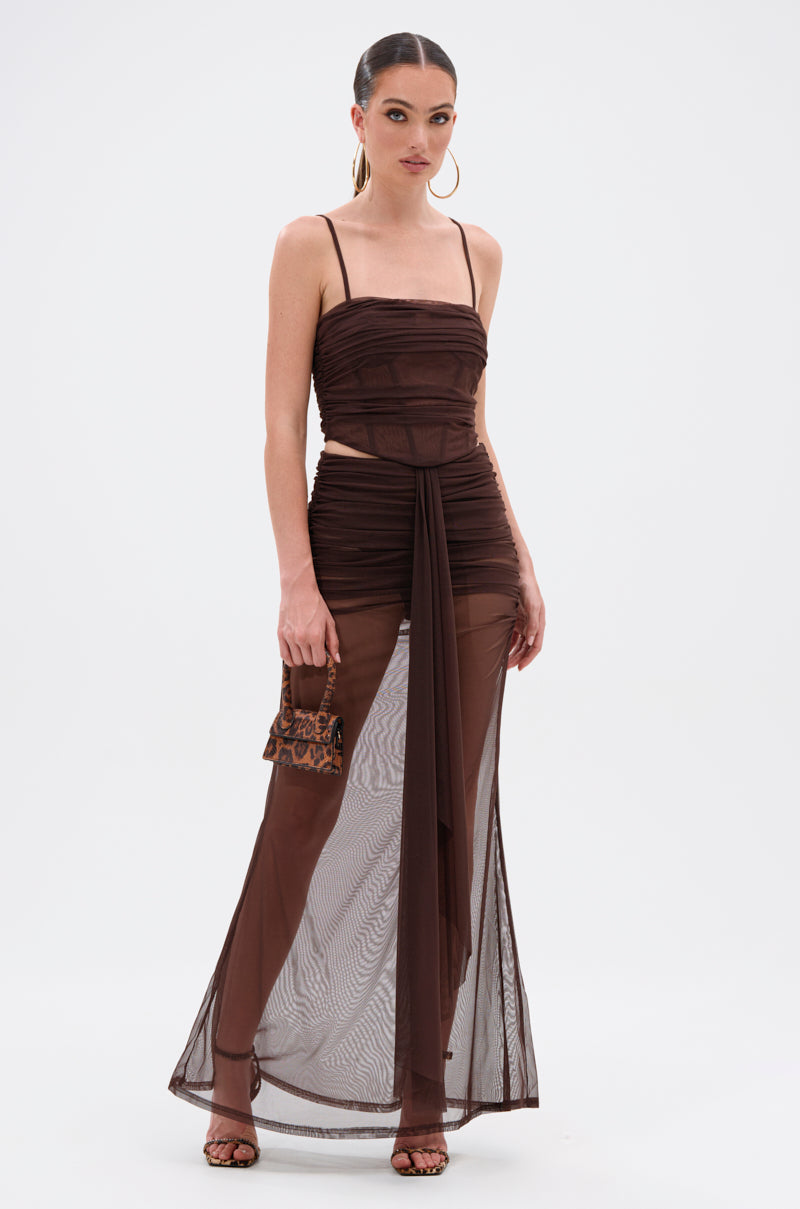 A woman wears the SHAKE IT FOR ME GIRL MESH MAXI SKIRT, a sheer, dark brown maxi skirt with ruched details and a draped front tie, paired with a leopard print purse, hoop earrings, and heeled sandals. 0