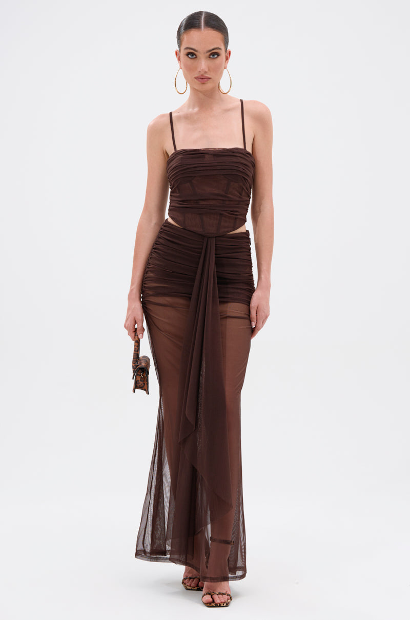 A woman in the SHAKE IT FOR ME GIRL MESH MAXI SKIRT—a sheer, dark brown maxi skirt gown with thin straps, ruched bodice, draped sash at the waist—holds a small purse and pairs it with large hoop earrings and heeled sandals. 5
