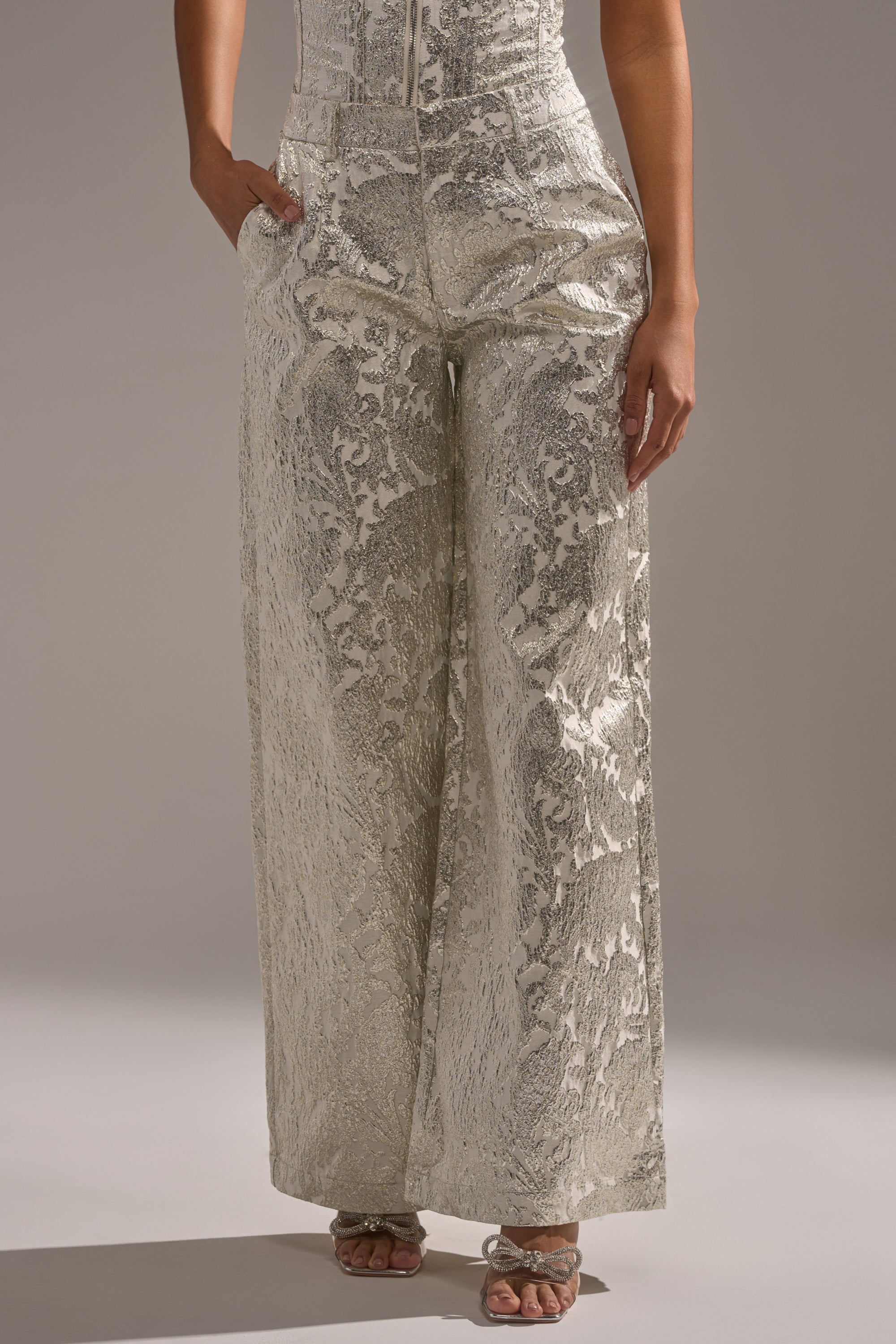 A person models the COCO BROCADE TROUSER—metallic, high-rise, wide-leg pants with a subtle floral pattern—perfect for holiday parties. They rest their hands in the pockets and wear open-toed heeled sandals against a neutral gray background. 0