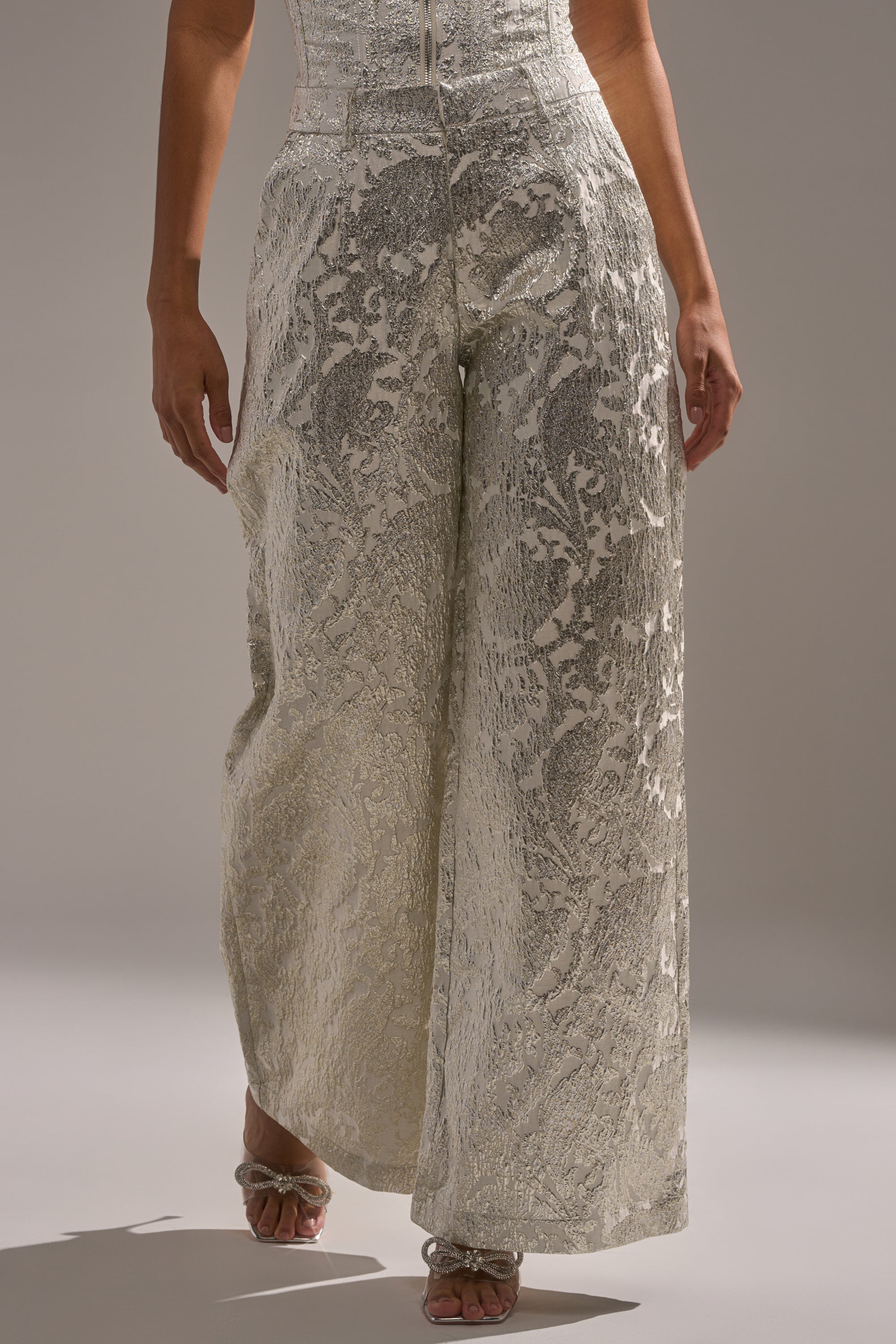 A person wears the COCO BROCADE TROUSER—high-rise wide-leg pants in metallic floral brocade—for a striking holiday party look, paired with open-toe heeled sandals and shown from the waist down. 1