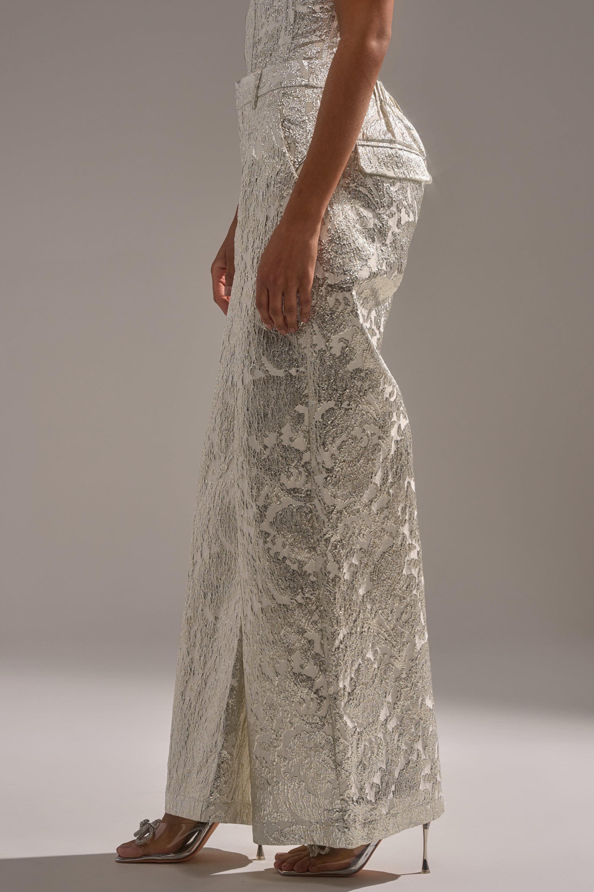 Wearing the COCO BROCADE TROUSER in textured metallic silver with a fitted waist and flared leg, paired with silver open-toe high heels. Only the lower half is visible against a neutral background—an ideal holiday party look. 2