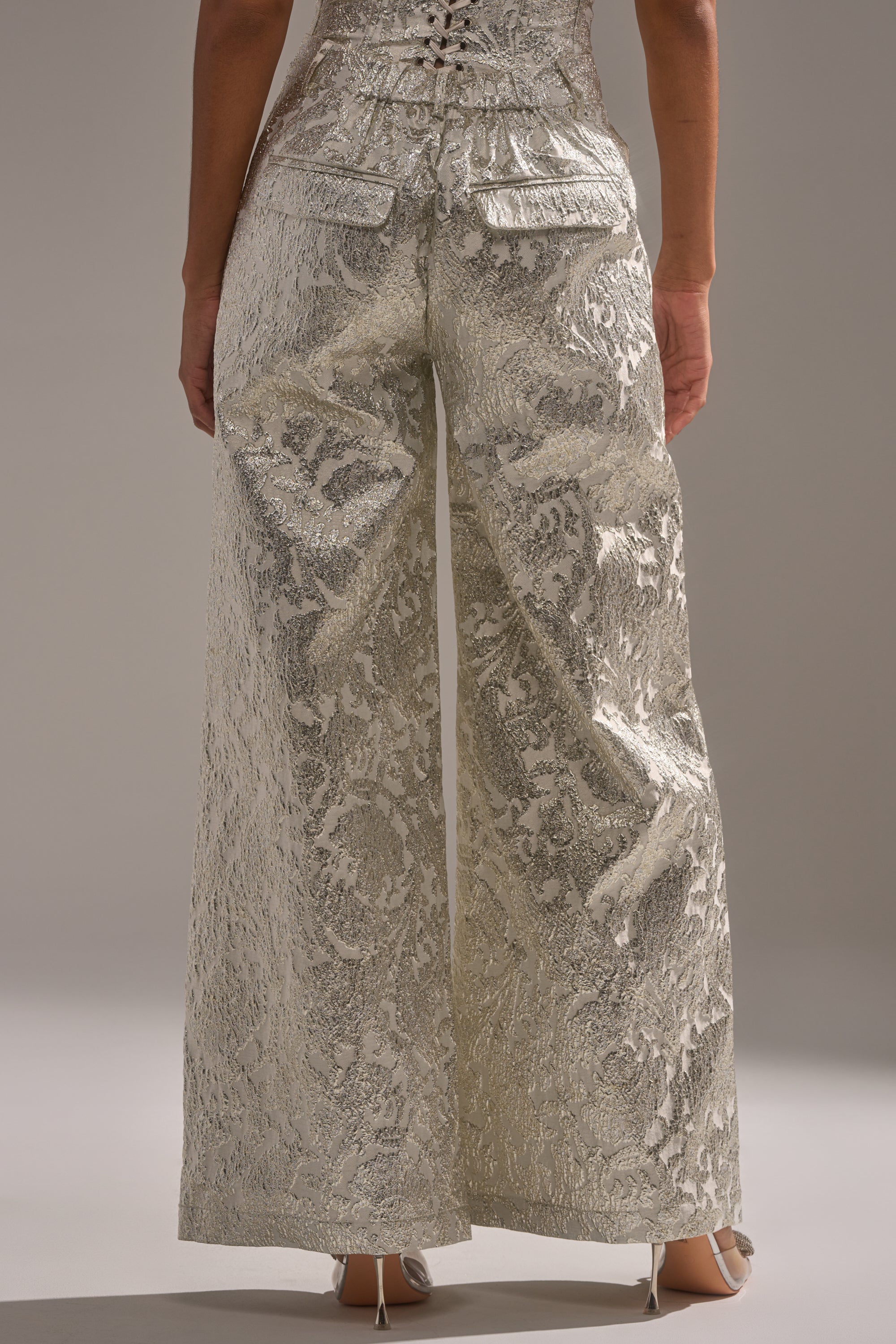 A person stands facing away, wearing the COCO BROCADE TROUSER—high-rise, wide-leg pants with a shimmery metallic brocade pattern. Only the lower torso and legs are shown, emphasizing the textured fabric and flap back pockets. Perfect for holiday parties. 3