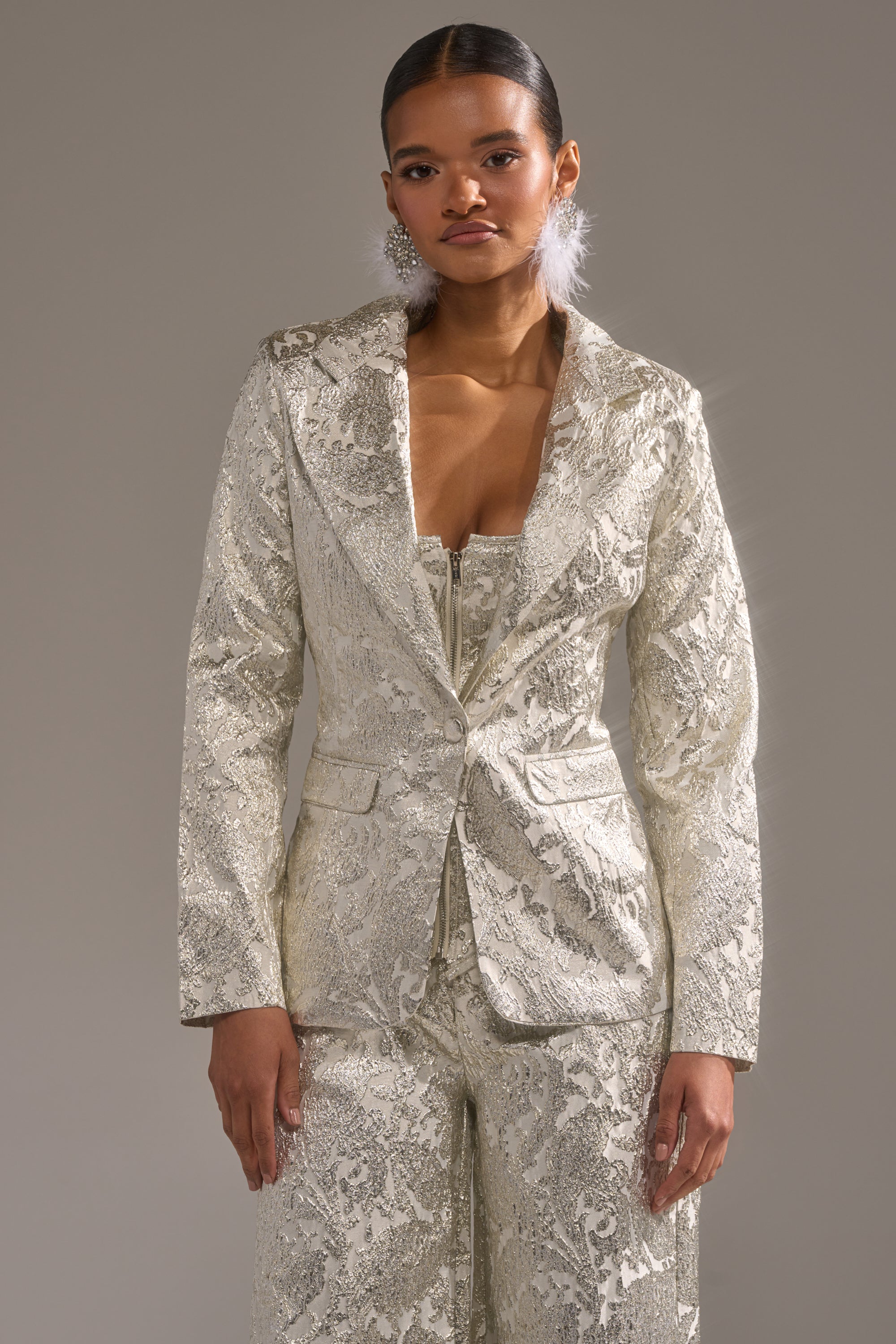 A woman with sleek, pulled-back hair and large white earrings stands against a neutral background wearing the COCO BROCADE BLAZER, a shiny, textured silver jacket paired with matching pants. 2
