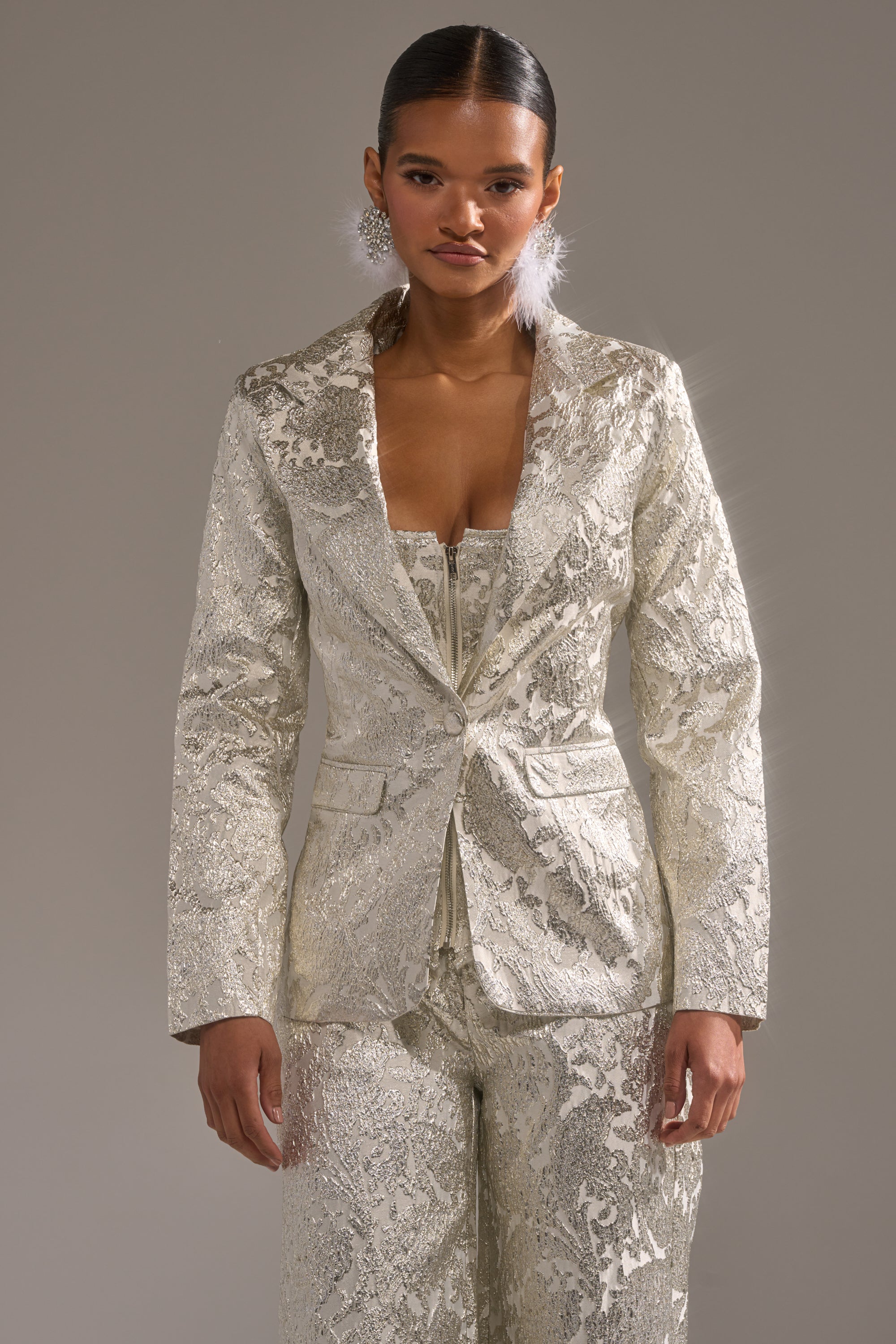 A woman poses against a plain gray background wearing the COCO BROCADE BLAZER, a shimmering textured silver longline pantsuit with a deep neckline. She completes the look with slicked-back hair and statement earrings. 0