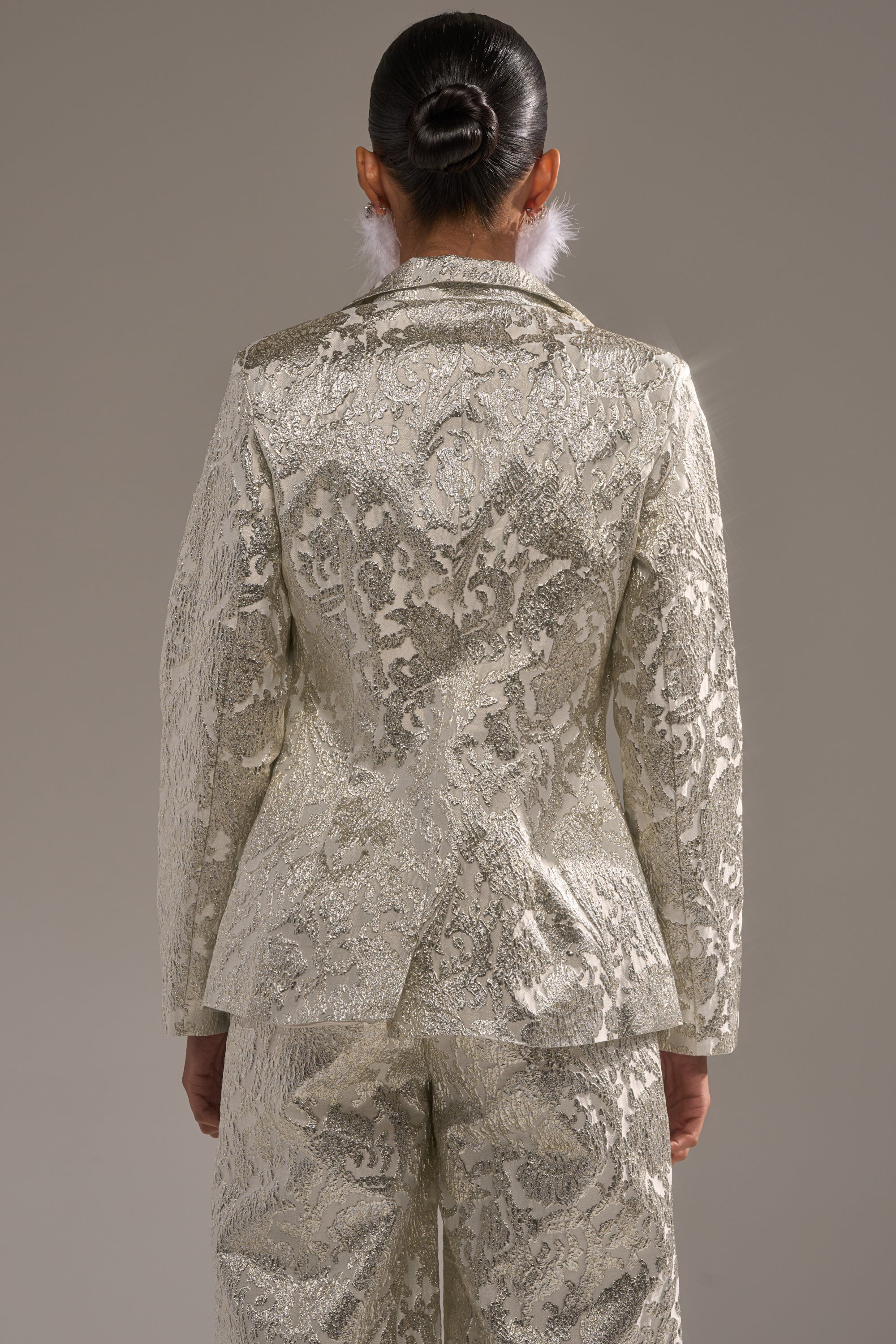 A person with dark hair in a neat bun wears the COCO BROCADE BLAZER and a matching cream and silver suit, seen from the back against a plain gray background. 4