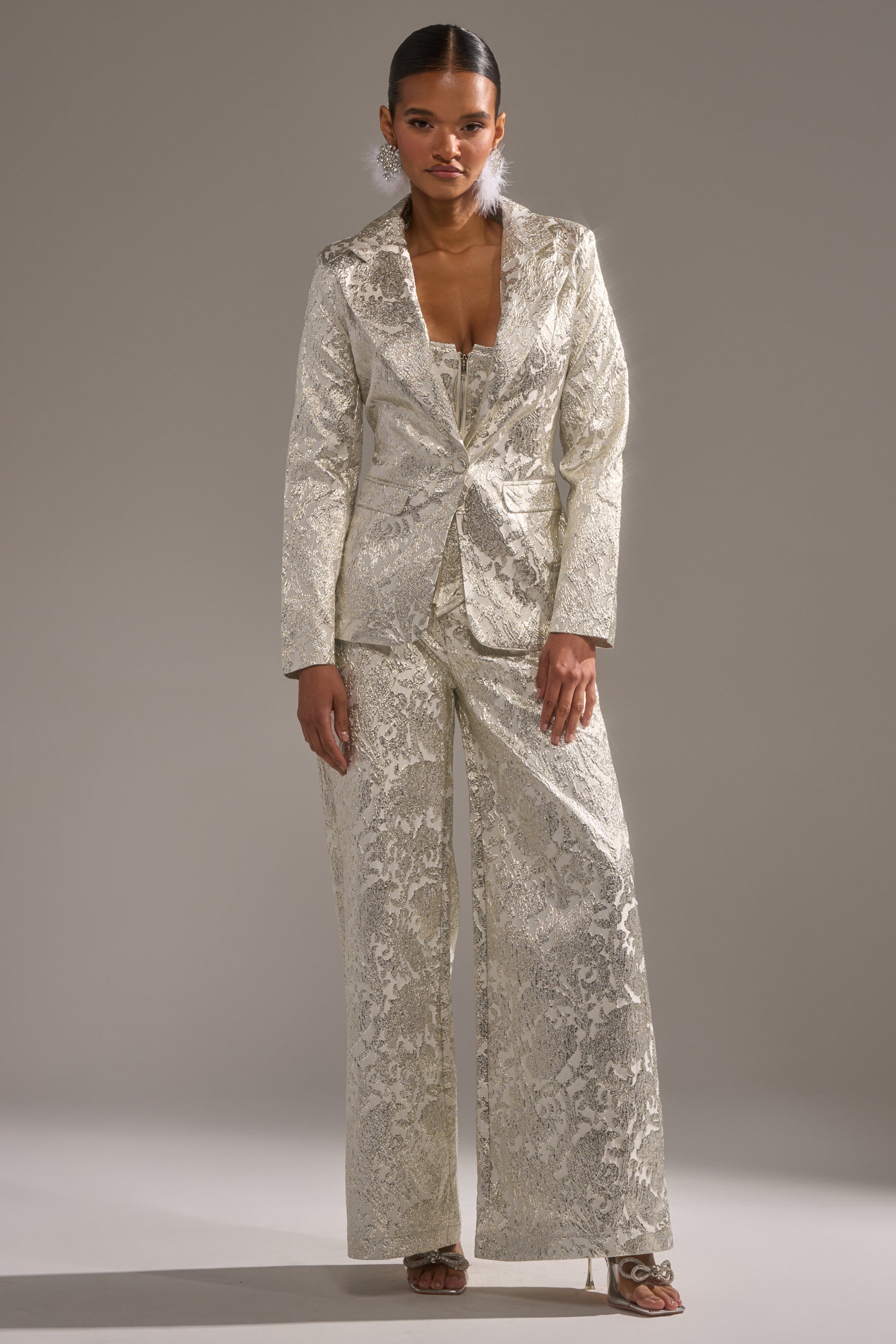 A woman faces forward in the COCO BROCADE BLAZER, an ivory metallic brocade jacket with wide-leg trousers. Her hair is pulled back, and she wears heeled sandals against a simple neutral background. 5