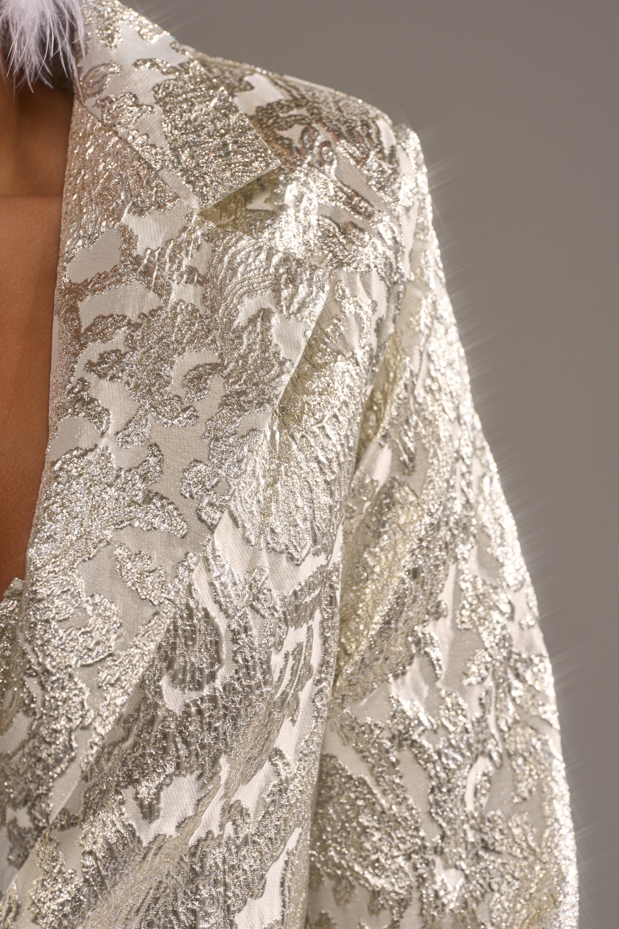 Close-up of a person wearing the COCO BROCADE BLAZER, a metallic gold and cream longline blazer with a textured floral pattern, set against a plain gray background. 6