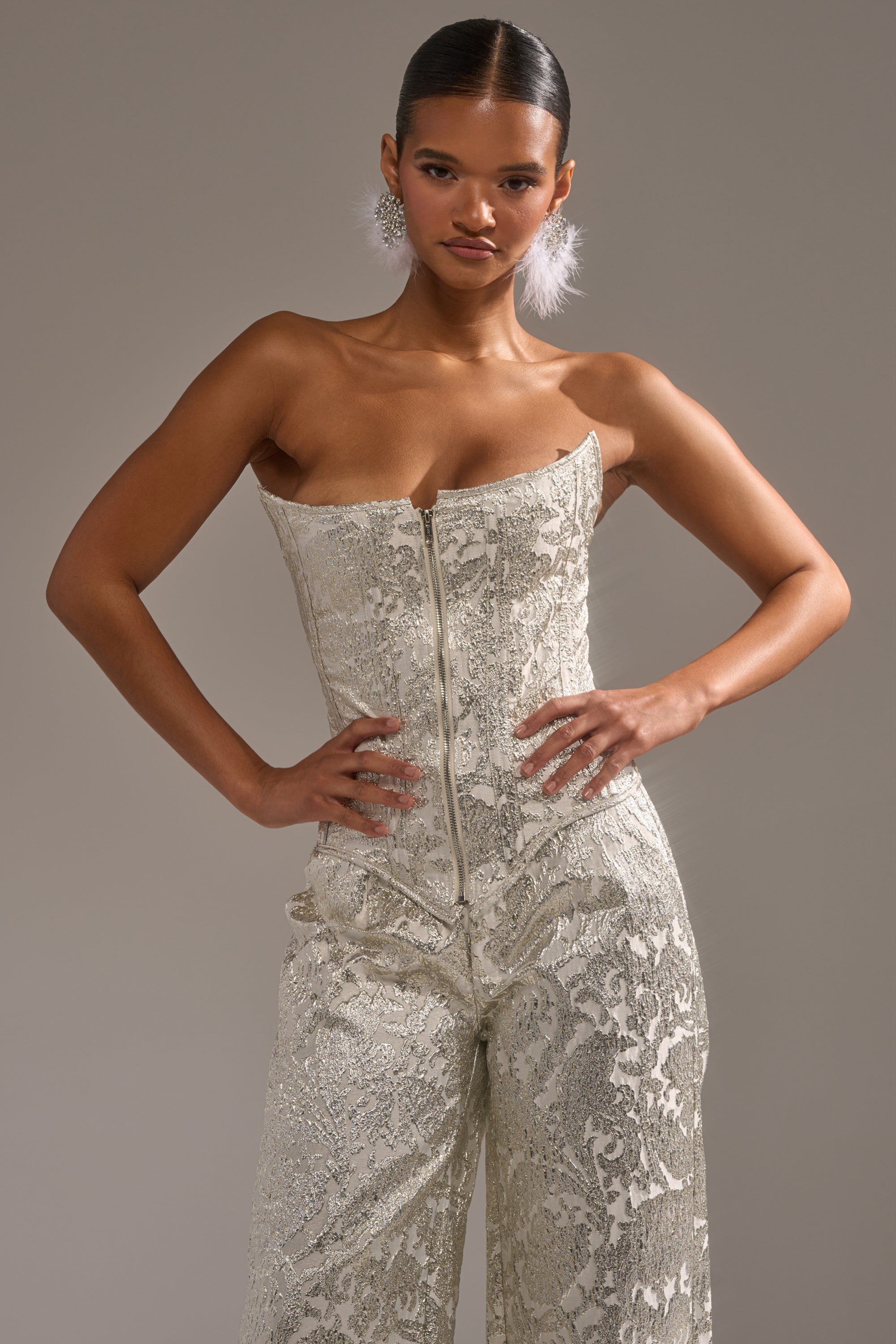 A woman poses confidently in the COCO BROCADE CORSET, a strapless metallic silver jumpsuit with a front zipper. She stands with her hands on her hips, slicked-back hair, feathered earrings, and a neutral background. 1