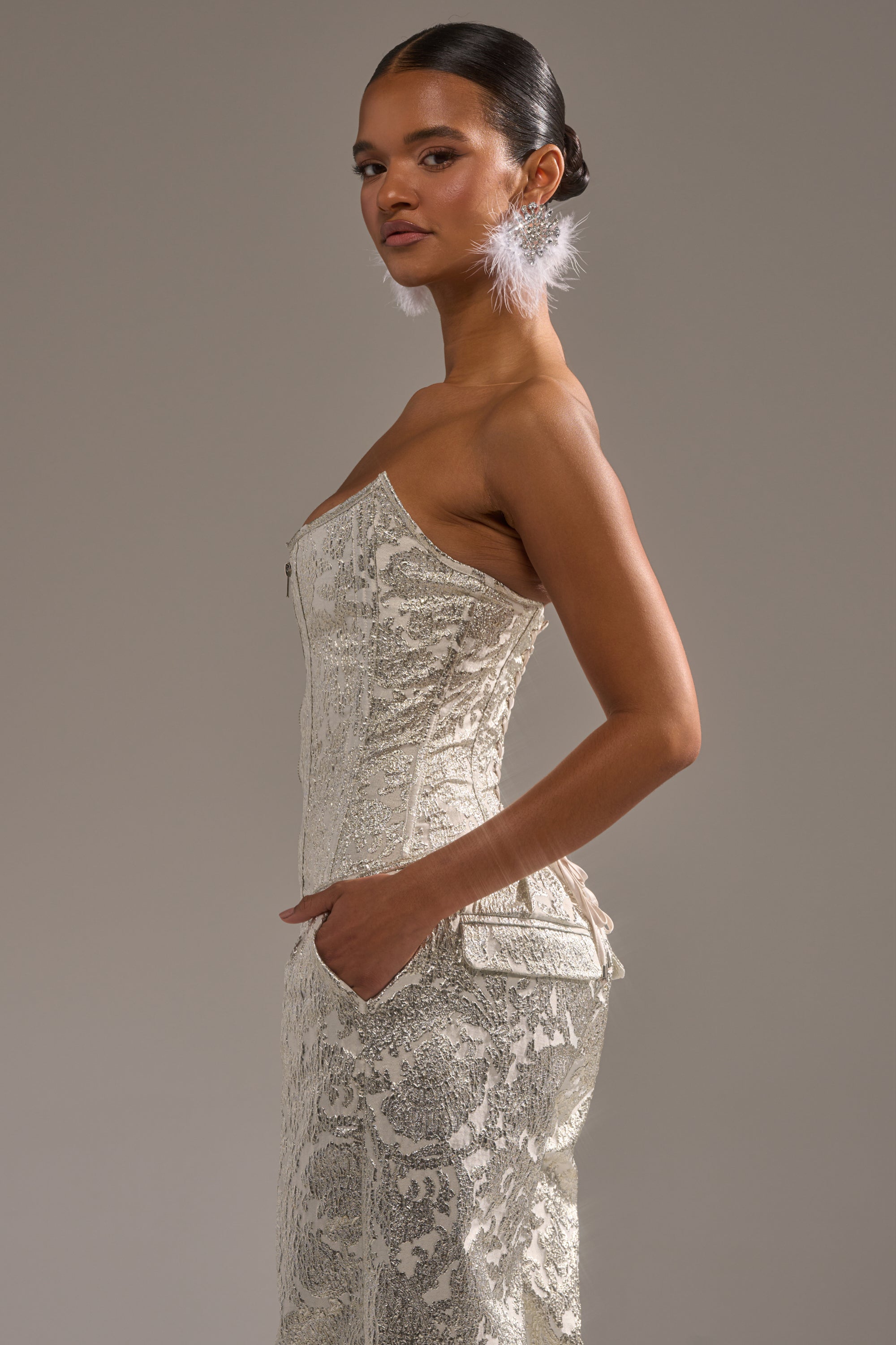A woman in profile wears the COCO BROCADE CORSET cream lace dress, her hair styled in a low bun. She poses with hands in pockets and large white feather earrings, set against a plain gray background. 2