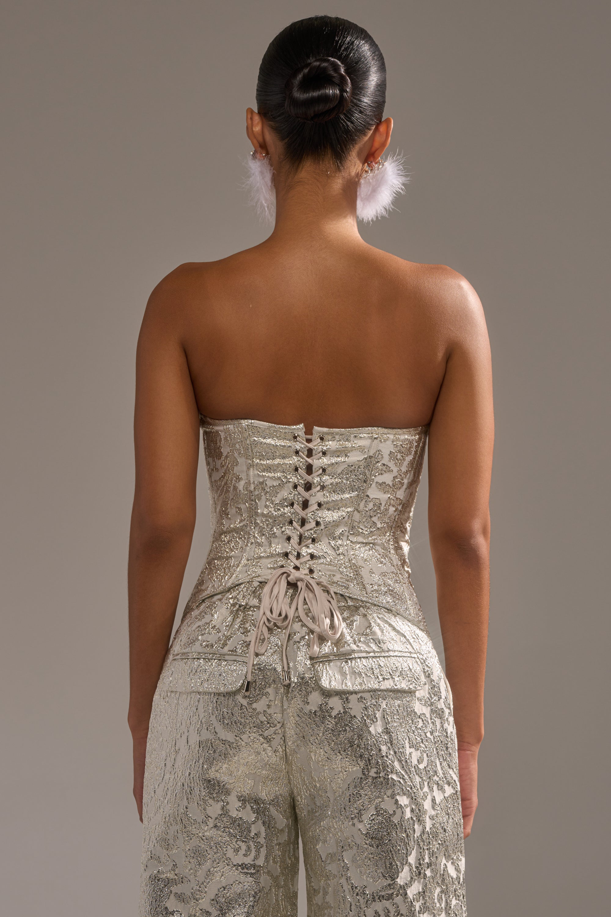 A woman with dark hair in a bun wears the COCO BROCADE CORSET, a strapless metallic silver piece laced up with ribbon, paired with matching pants and feather earrings. She is seen from the back against a plain gray background. 3