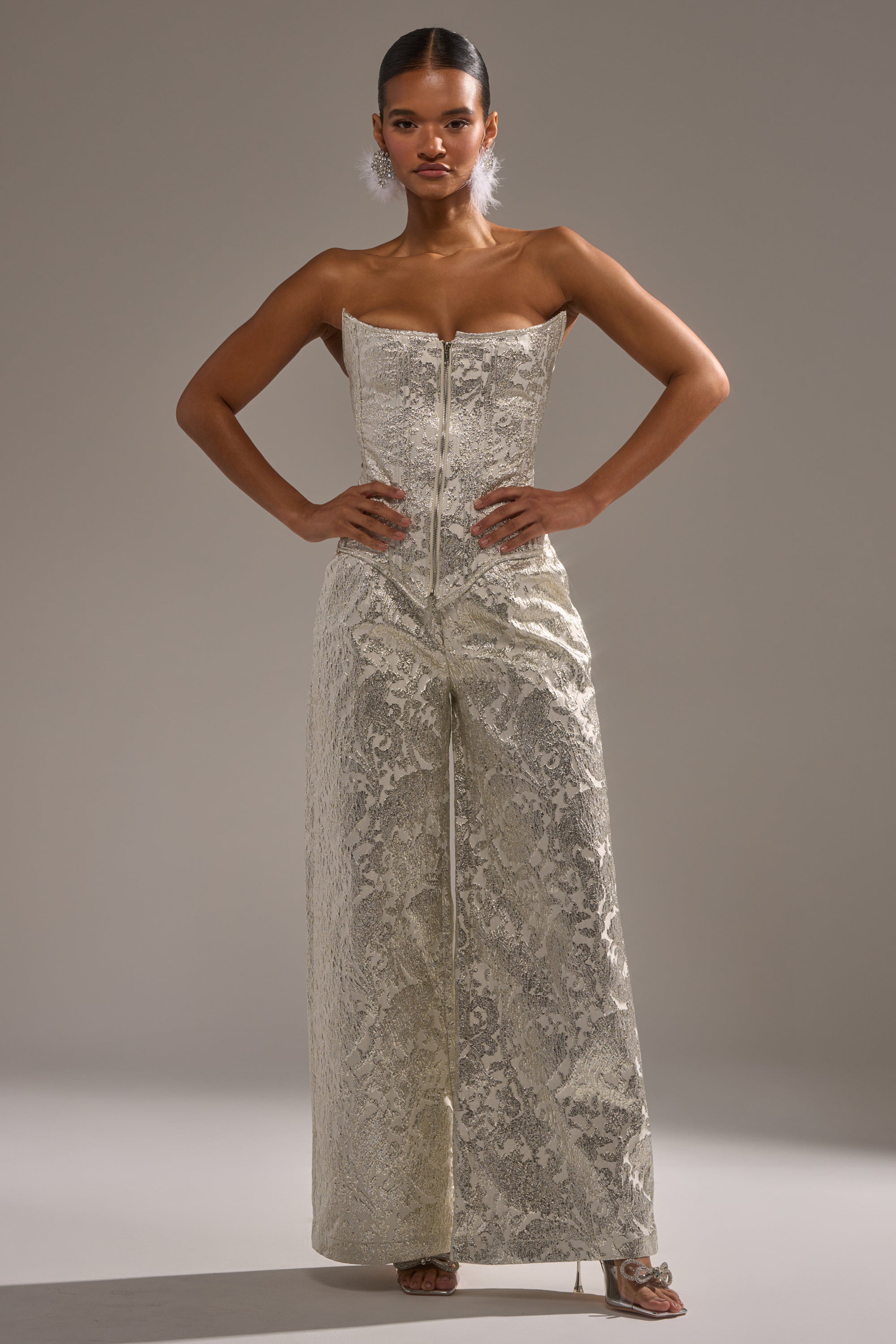 A woman stands confidently with hands on hips, wearing the COCO BROCADE CORSET—a metallic silver jumpsuit with a front zipper and wide legs. A neutral background emphasizes her striking outfit and poised stance. 5