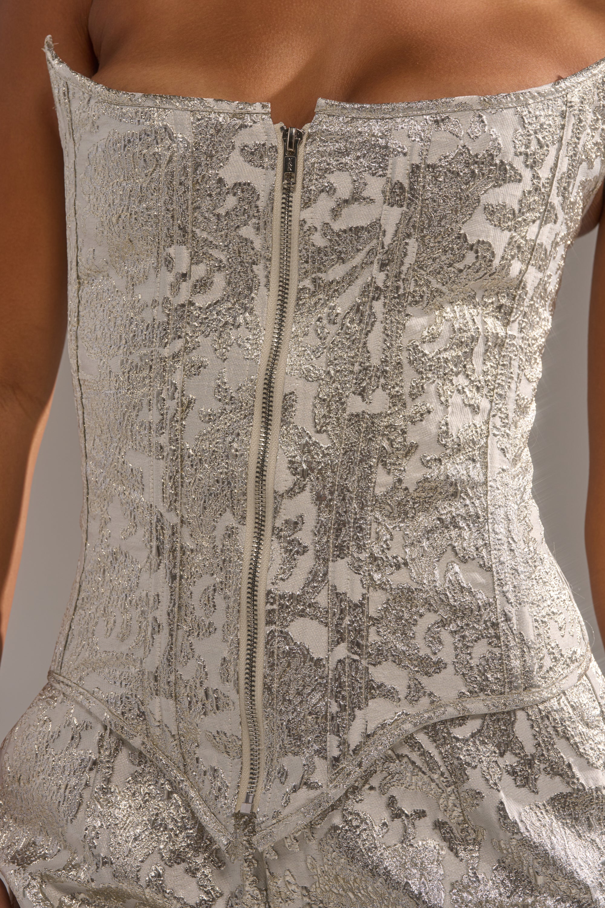Close-up of a person wearing the COCO BROCADE CORSET, a strapless metallic silver corset with a front zipper and shiny, intricate floral brocade for an elegant look. 6
