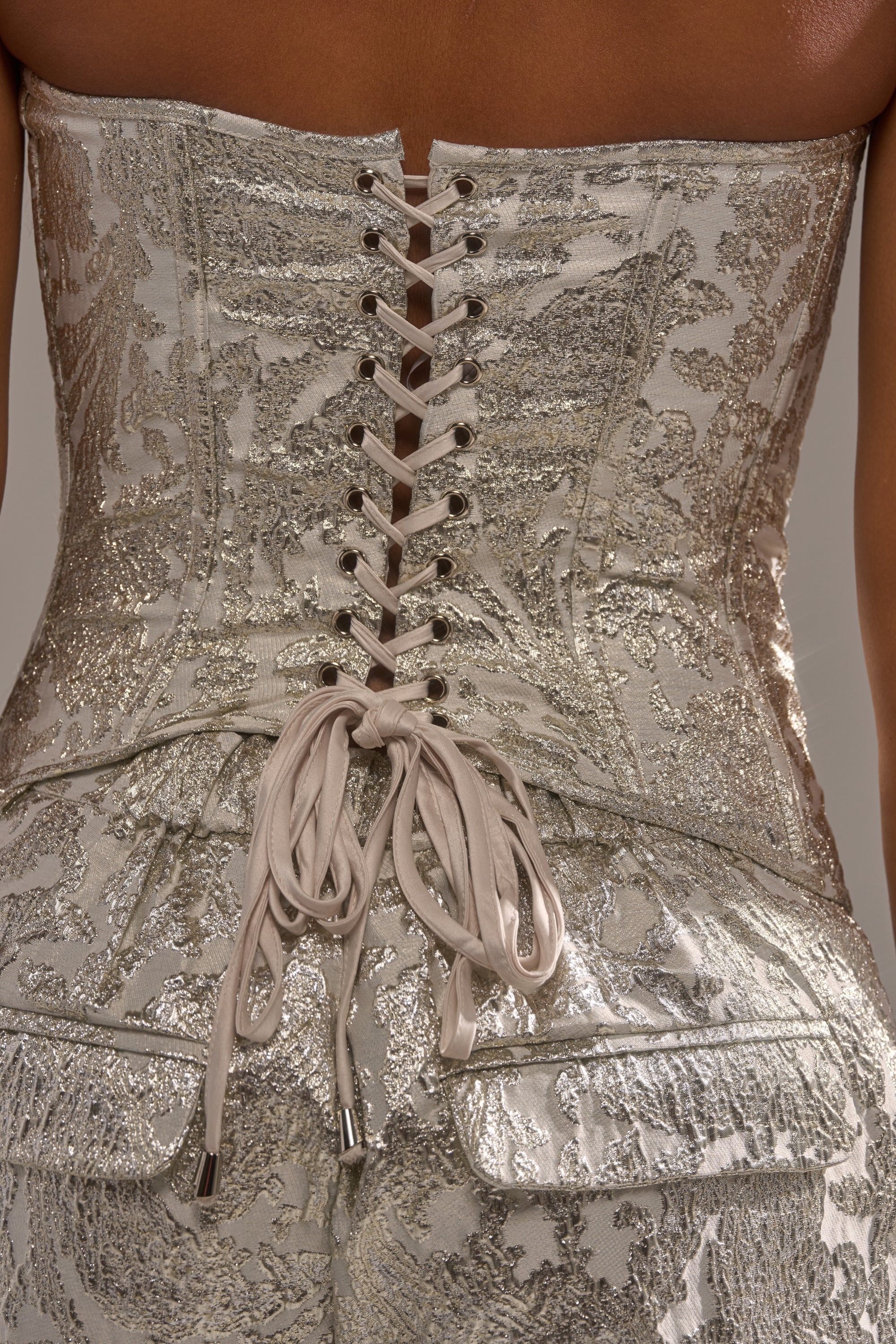 Close-up of the COCO BROCADE CORSET, a strapless metallic silver corset with floral brocade fabric and intricate lace-up back tied in a bow, showcasing its shiny textured finish. 7