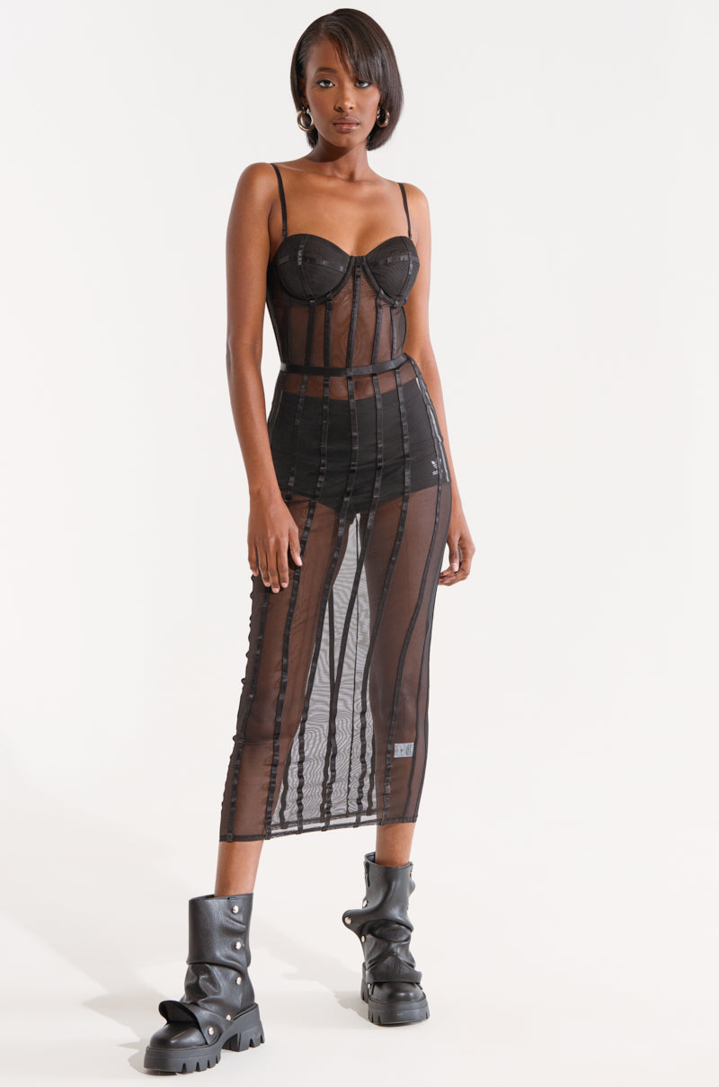 A woman stands against a plain white background in the SHE'S SNATCHED MESH MIDI DRESS, showcasing its cage-like grid pattern and bodycon silhouette. The black sheer dress, complete with a fitted bodice and structured cups, subtly reveals black underwear underneath. She complements her look with combat boots and stud earrings. 0