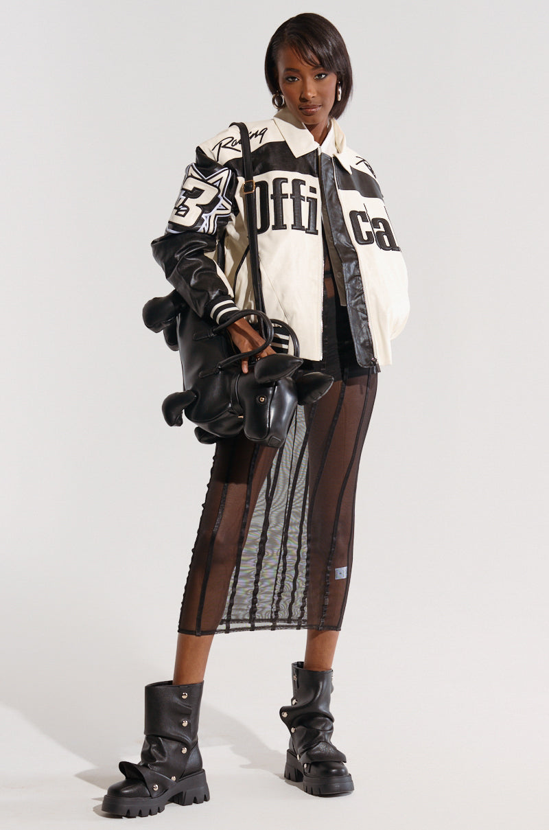 A fashion model confidently poses against a plain background, sporting the chic "SHE'S SNATCHED MESH MIDI DRESS," which features a see-through, vertically striped skirt with a cage-like grid patterning. She completes her stylish ensemble with an oversized white and black jacket adorned with bold text and pairs it with chunky black boots. A sleek black handbag adds the finishing touch to her look. 1