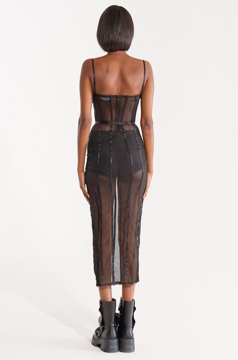 A person stands with their back to the camera, showcasing the SHE'S SNATCHED MESH MIDI DRESS. This black, sheer dress features vertical stripes and a bodycon silhouette, complemented by black chunky boots. They wear their hair in a short, sleek bob. The dress boasts spaghetti straps, a snug fit, and falls below the knees. 3