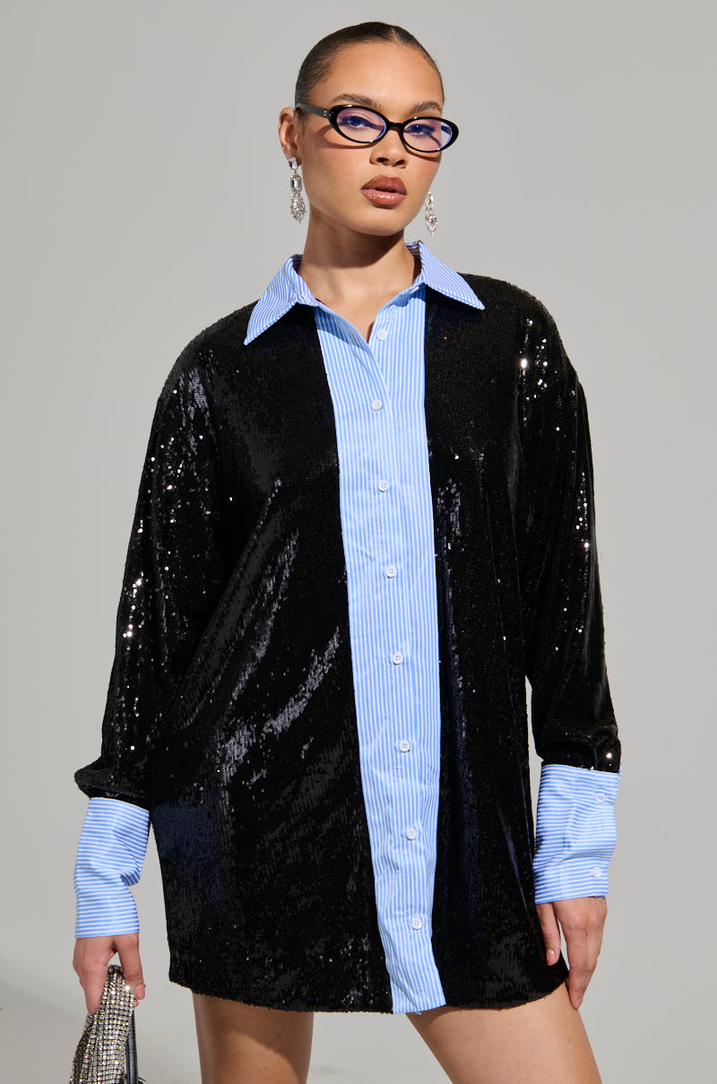 A person is wearing the CYRUS SEQUIN POPLIN SHIRT DRESS, featuring an oversized fit with a shiny black finish and a blue and white striped collar and cuffs. They have short hair, wear glasses, and are holding a small bag, set against a plain gray background.